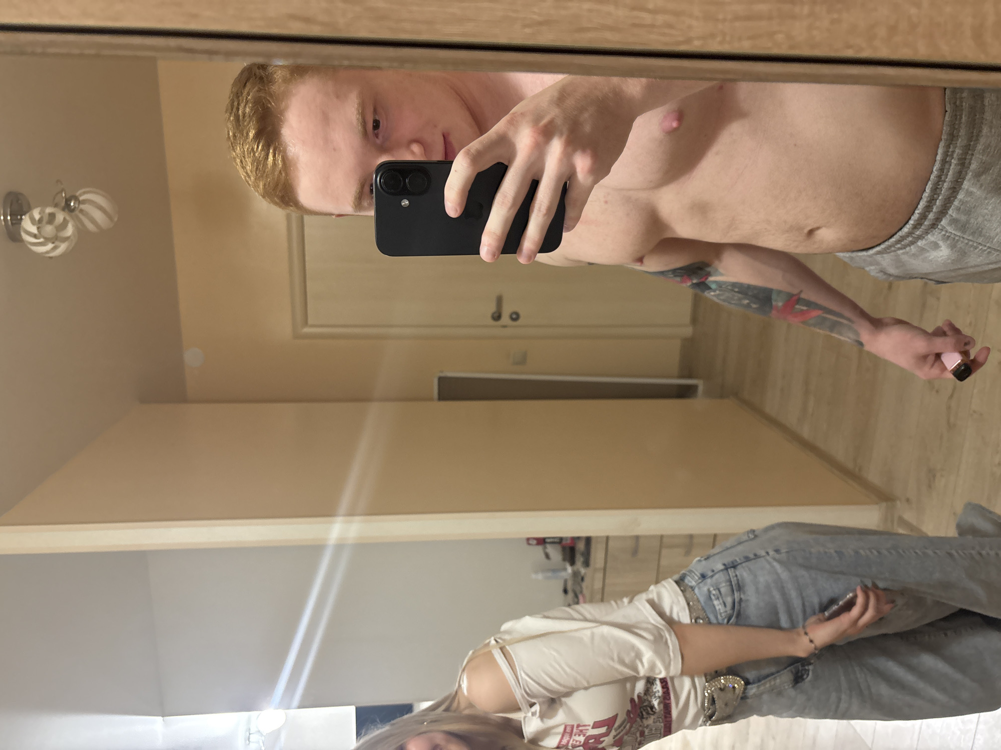 A mirror selfie captures a shirtless fair-skinned man with red hair holding a black smartphone. A partial view of a woman with blonde hair wearing a white shirt and jeans is visible in the mirror's reflection. The scene appears to be indoors, possibly a hallway or living space. The location is Chaikovsky, Russia. A wall fixture with three circular lights is visible on the wall.