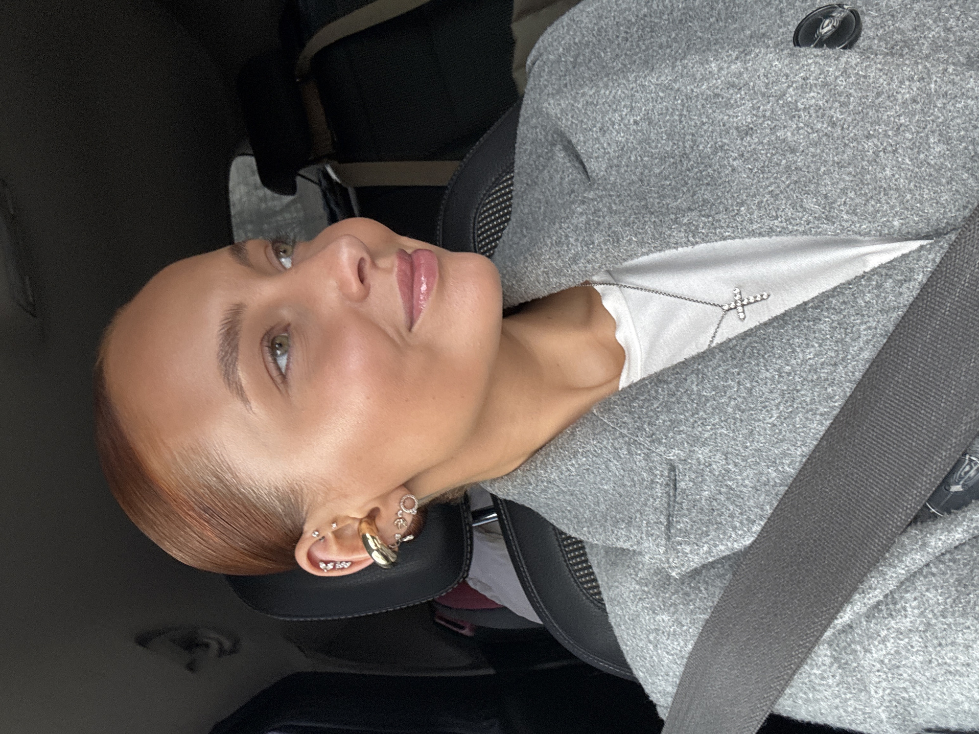 A portrait-oriented image shows a young woman from the chest up, rotated 90 degrees clockwise, so her head is on the left side of the frame, looking towards the top right corner. She has fair skin, light brown hair pulled back, and light-colored eyes, possibly hazel. Her lips are glossy, and her makeup appears natural. She wears a light grey textured coat over a white top, accessorized with a silver cross pendant necklace. Her visible left ear features multiple piercings, including several studs, a gold-colored helix cuff, and a dangling earring.
She is seated in a car, with a black seatbelt diagonal across her chest and shoulder, and a black car seat headrest visible behind her. The background is dark and blurry, showing parts of the car's interior. Her expression is serene and contemplative, with a subtle upward gaze and a slight, pleasant smile. The lighting suggests it is daytime. The scene takes place in Chaikovsky, Russia. There is no visible text in the image.