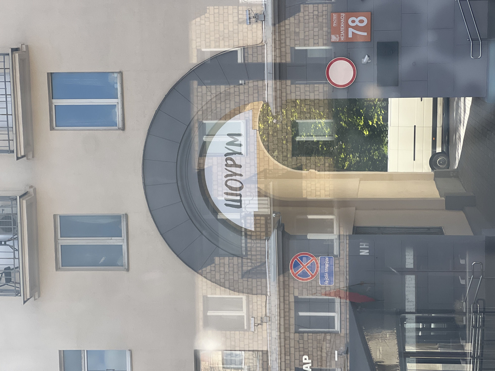 This image captures a reflection on a window, showing a street scene in Minsk, Belarus. The primary focus is a building with an arched entryway. On the arch, the word "ШОУРУМ" (Showroom) is visible in large white letters.
To the left of the arch, a building with brick facade and windows is reflected. A red flag hangs from a pole near a blue sign with a red circle and a diagonal line, indicating "no stopping," and text below it in Belarusian. Another sign with the letters "AP" is partially visible. Further to the left, the letters "NH" are visible on a dark facade.
To the right of the arch, another building is reflected, also with brickwork and windows. A red circular sign and a rectangular sign with a coat of arms are mounted on the wall. The coat of arms is accompanied by text that appears to be "ПРАСПЕКТ НEЗАЛEЖНАСЦI" (Independence Avenue) and the number "78" on an orange background. A dark rectangular plaque is mounted below these signs.
Through the archway, a white van is parked on a paved area. Green foliage from trees is visible behind the van. The overall scene suggests a commercial or public area.