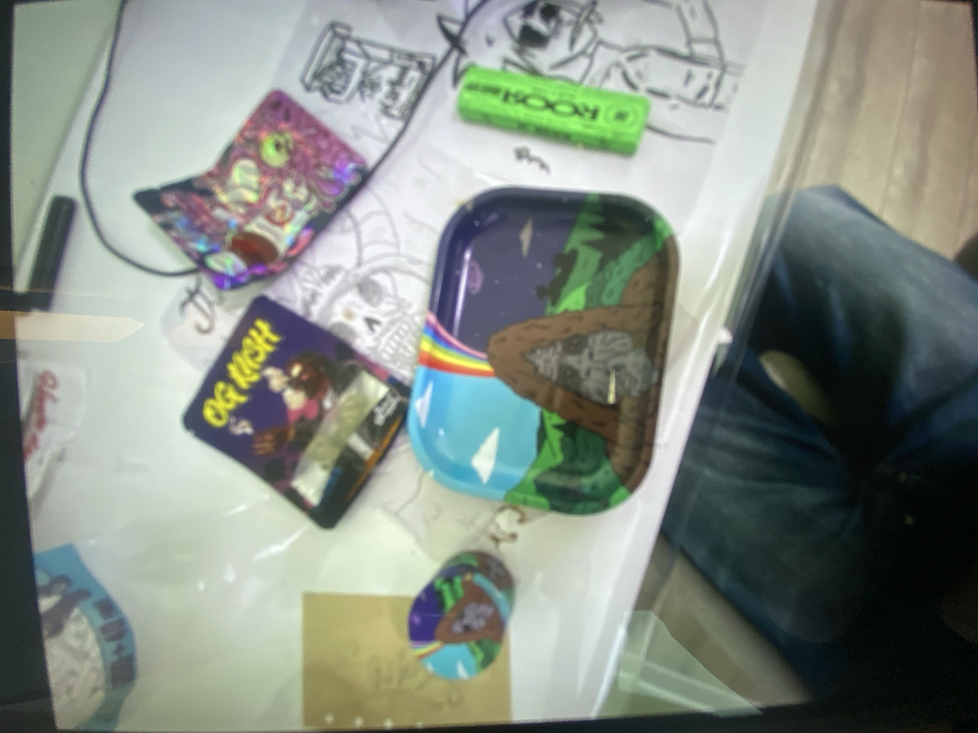 The scene presents a variety of objects on a white surface, likely a table. Items include several small, rectangular packages with colorful and cartoonish designs, such as one labeled "OG Kush". There is also a small, green rectangular object with the word "BOOM" visible. A small tray with a design featuring mountains, a starry sky, and a rainbow bridge is present, along with a smaller circular object with a similar design. In the background, partial view of a person wearing jeans is discernible.
The location of the scene is unclear in the city of Plovdiv, Bulgaria.
