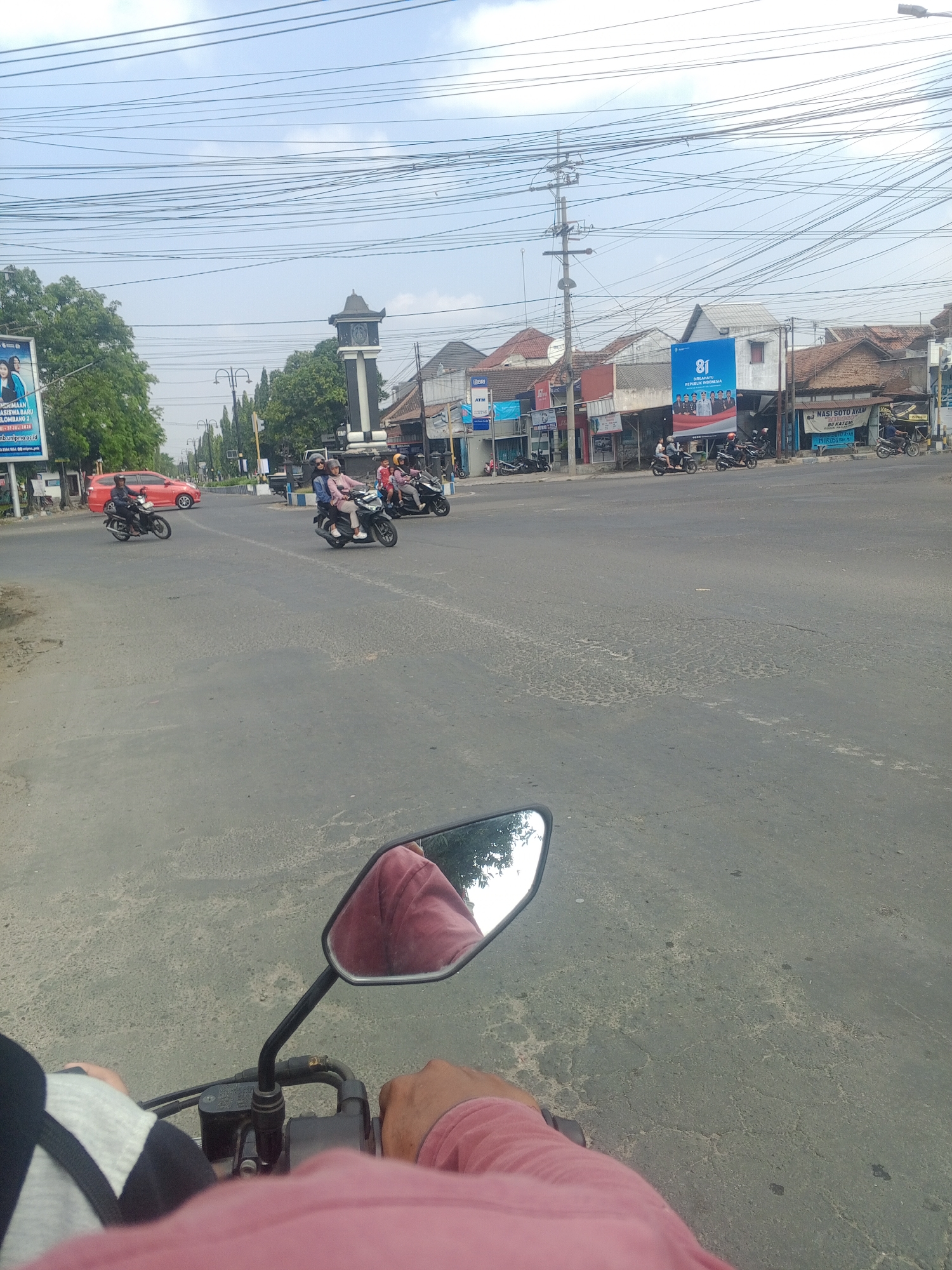 The image captures a street scene in Mangunsuman, Indonesia, with a first-person perspective from a motorcycle. The road is paved and shows signs of wear and tear, with visible cracks and patches. In the mid-ground, a traffic intersection is active with several motorcycles carrying passengers. To the left, a red car is in motion. Further in the background, buildings line the street, some featuring signage. A prominent clock tower stands near the intersection, indicating a town center or landmark. Overhead, a dense network of power lines crisscrosses the sky, typical of urban environments. The sky is partly cloudy. The overall impression is of a bustling, everyday scene in an Indonesian town.
