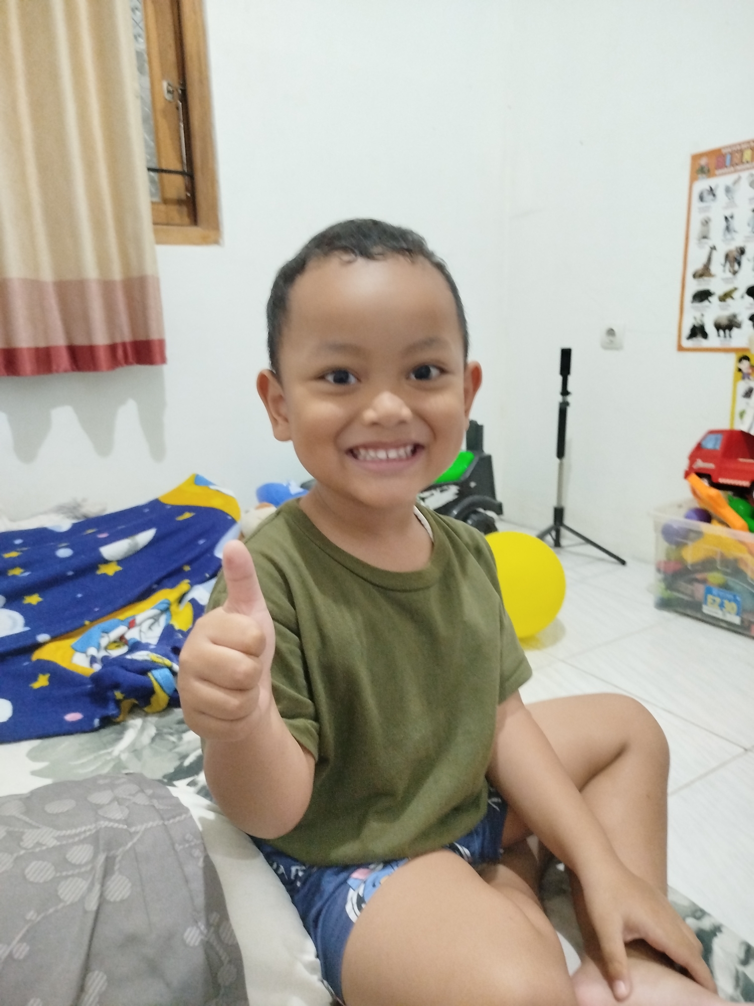 A young Indonesian boy with dark hair and brown skin is sitting on a bed in a room, giving a thumbs-up gesture. He is wearing a short-sleeved olive green t-shirt and blue shorts with a white pattern. The bed has a blue blanket with yellow stars and cartoon characters. In the background, a yellow balloon is visible, along with toys and a poster of animals on the wall. A curtain hangs over a window to the left, and a tripod is set up behind the boy. The scene appears to be indoors, possibly a bedroom, in Jeruksawit, Indonesia.