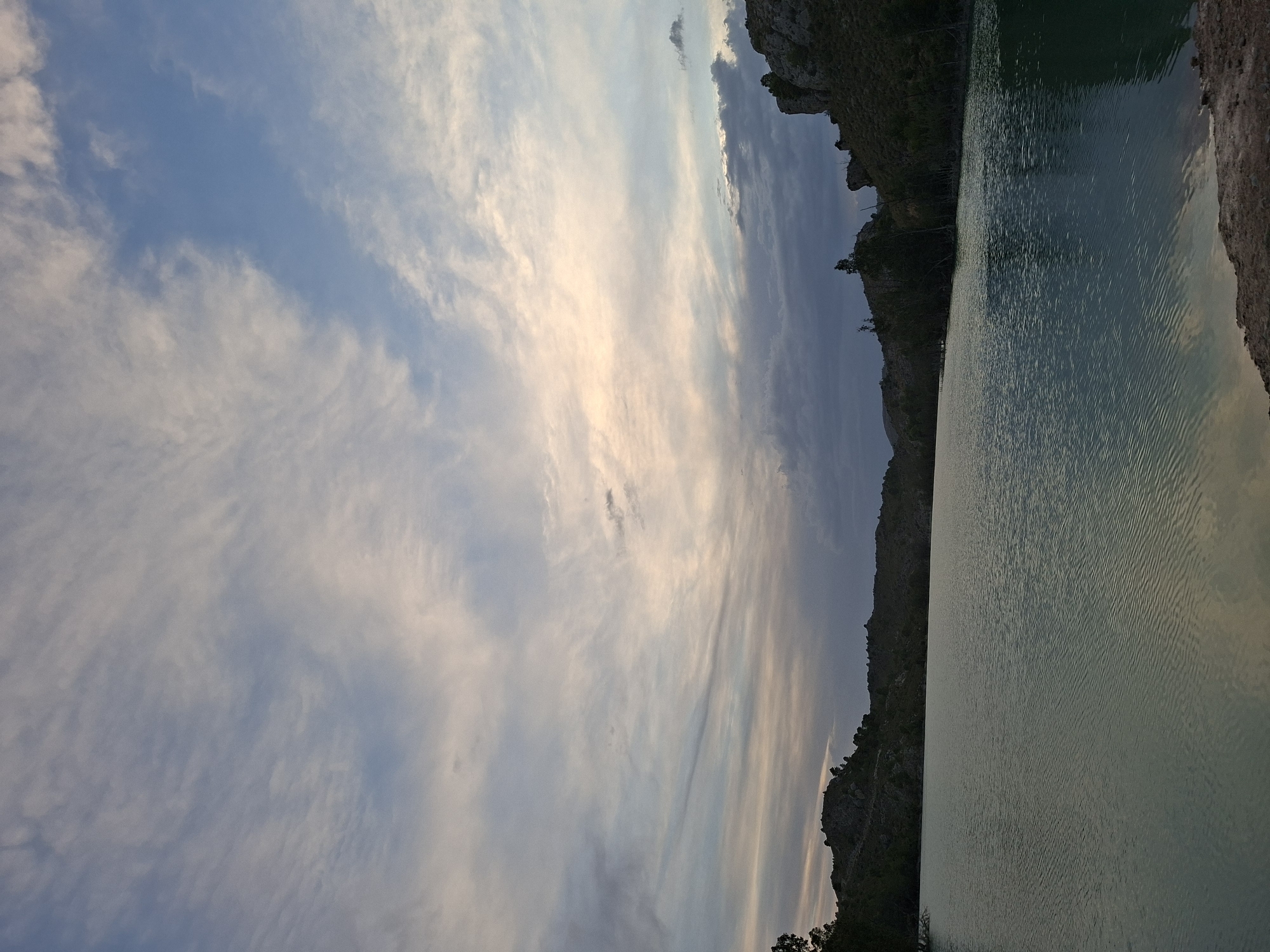 The image depicts a tranquil body of water, possibly a reservoir or lake, situated at the foot of rolling, arid hills. The sky above is a canvas of pastel hues, with wispy clouds stretching across the horizon, transitioning from pale blue to soft pinks and oranges, suggesting either dawn or dusk. The water's surface is calm, with gentle ripples reflecting the colors of the sky and the surrounding landscape. Dense vegetation, characterized by scattered trees and shrubs, clings to the slopes of the hills, adding texture to the scene. The overall impression is one of natural serenity, with the landscape bathed in the soft, diffused light of either early morning or late evening. The location is the reservoir of Algeciras, near Mula, Spain. There are no people or animals visible in the image.