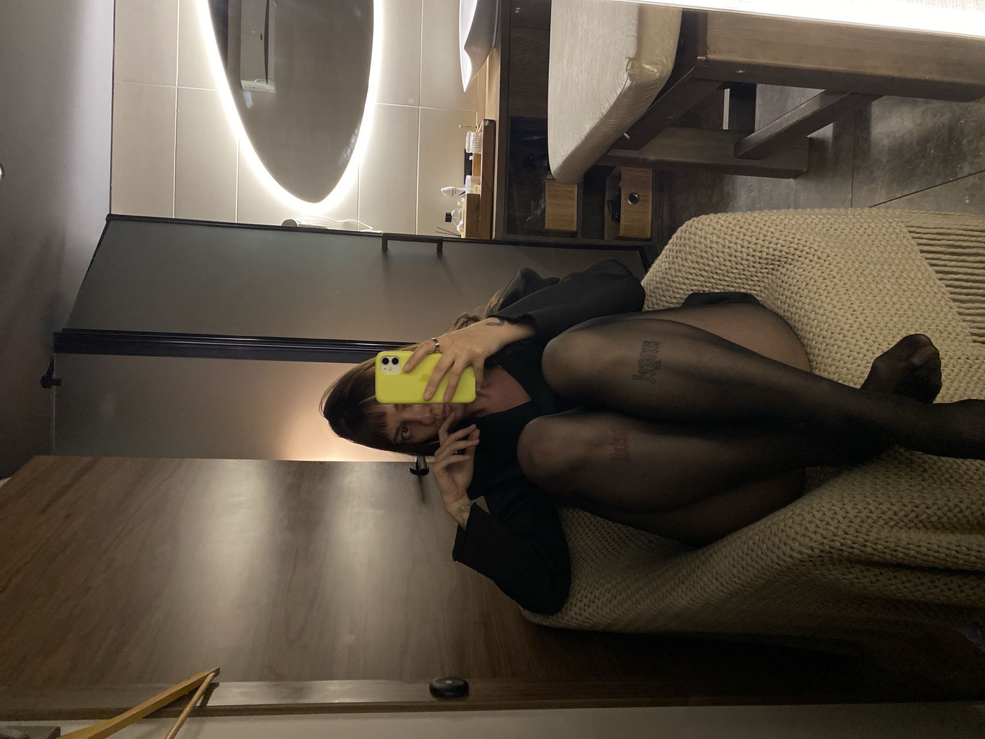 A woman is captured in a mirror selfie within what appears to be a modern hotel room, spa, or private treatment room in Votkinsk, Russia. She is seated in a light-colored, textured armchair draped with a matching knitted blanket featuring fringes, her legs crossed. She wears a black v-neck top or blazer and sheer black tights. On her left thigh, a red script tattoo is visible, while on her right thigh, a dark tattoo in a Gothic-style script font is present. She holds a bright yellow smartphone, likely an iPhone with multiple rear cameras, in her right hand, taking the photograph of her reflection. Her dark hair features bangs, and she has rings on her right hand.
The reflection in the mirror reveals a contemporary interior. To the woman's right, a light-tiled wall features a wooden counter with a white basin sink and a wooden tray holding various personal care products, bottles, and jars. Below the counter, open wooden shelves contain woven storage baskets. Further to the right, a light-cushioned massage or examination table is visible. Above the counter, a large oval mirror with an integrated LED light border provides illumination. A white air conditioning unit is mounted on the wall above the vanity area, its reflection visible within the large oval mirror. To the woman's left, a wooden door with a dark handle and a light-colored wooden clothes hanger on a hook are visible. The floor is covered with dark gray tiles. The lighting is warm and ambient.