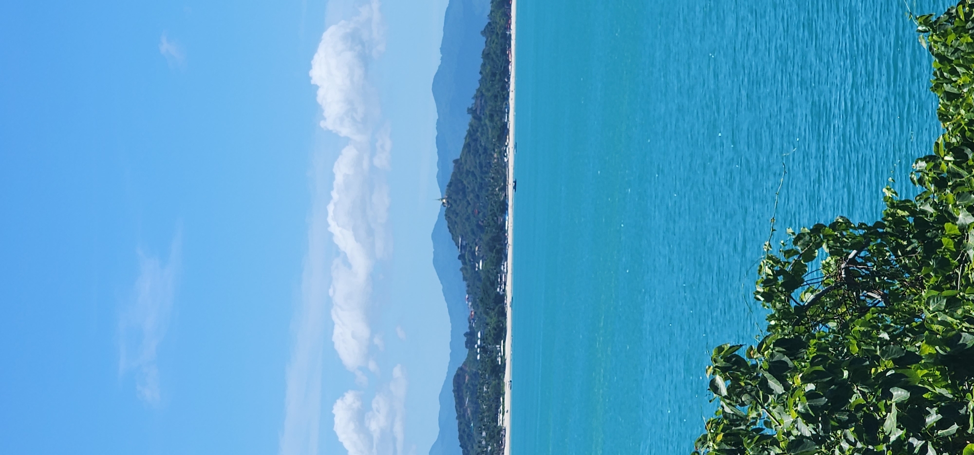 The image shows a panoramic view of a coastline in Baan Chaweng Noi, Thailand. In the foreground, lush green foliage is visible, partially obscuring the vibrant turquoise ocean. Beyond the water, a sandy beach stretches across the horizon, lined with palm trees and scattered buildings. A prominent green hill rises from the coastline, topped with a golden stupa, indicating a Buddhist temple. The background features rolling blue hills and a clear sky with scattered white clouds. Several small boats are visible on the water. The scene depicts a serene and tropical landscape, likely during daytime given the bright sunlight.