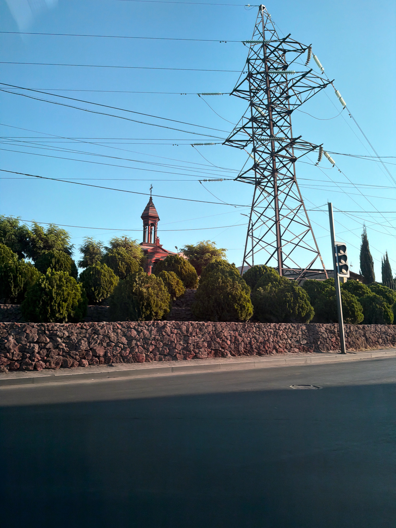 This is a ground-level, eye-level, medium-range, outdoor, wide-angle shot of a scene in Yerevan, Armenia. The scene is dominated by a large metal transmission tower with high-voltage power lines stretching across the sky. In the middle ground, a low, textured stone wall borders a raised area with lush green, manicured trees. Behind the trees, a red-brick building with a steeple and a cross is visible. A traffic light is positioned on the right side of the frame. The foreground is a dark asphalt road with a distinct shadow. The sky is clear and blue.