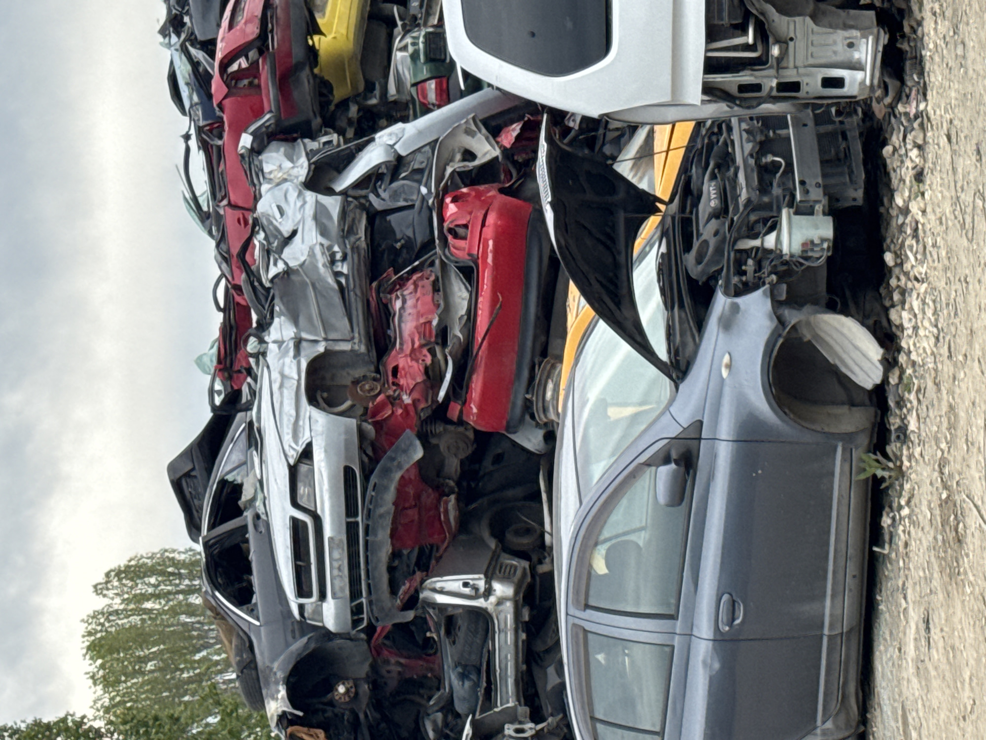 A vertical shot depicts a stack of damaged car parts, possibly at a junkyard in Varna, Bulgaria, or nearby area. The parts are stacked high, with a sky visible in the background. The parts are from various colored cars, including red, yellow, silver, white, and blue. There is a tree line visible at the bottom left. A gray concrete surface is present on the right side. The car parts appear to be crushed and broken.