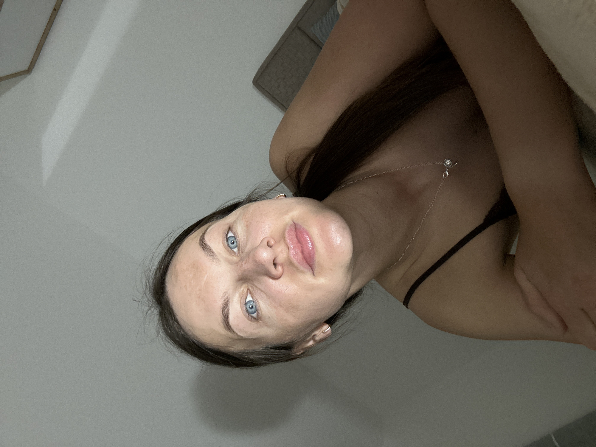 A woman with blue eyes and dark hair is lying down, likely on a bed, in an indoor setting. She is wearing a delicate silver necklace and a dark top. The background features a light grey wall and a woven grey headboard. The location, Budva, Montenegro, cannot be confirmed from the image.