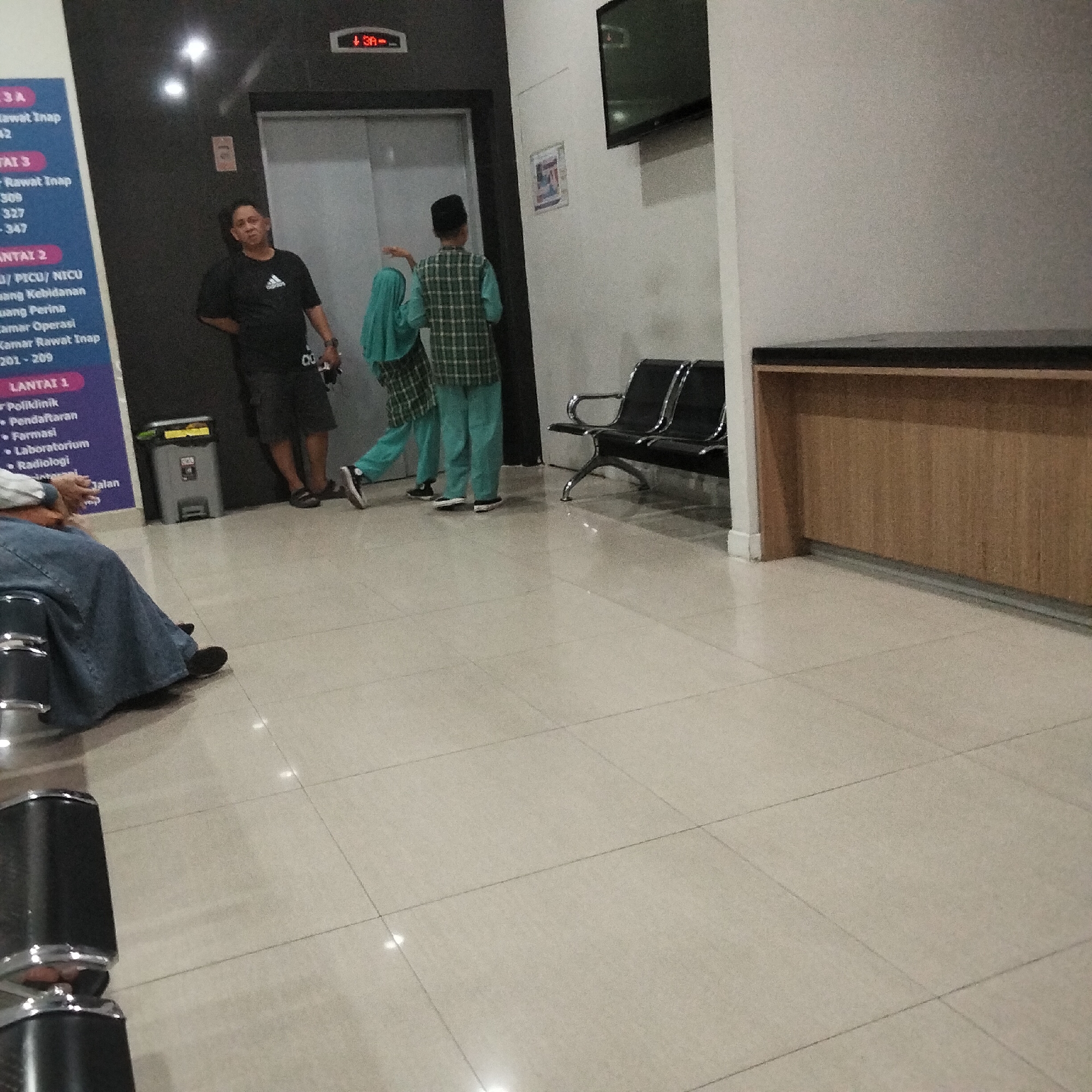 The image depicts an indoor waiting area with a man standing near an elevator. To the left, a blue sign lists floor levels and services like "Rawat Inap" (Inpatient Care), "PICU/NICU," "Kamar Operasi" (Operating Room), "Poliklinik" (Polyclinic), "Pendaftaran" (Registration), "Farmasi" (Pharmacy), and "Laboratorium" (Laboratory). Two individuals, possibly a woman and a child, are standing to the right of the elevator, one in a hijab and the other in a checkered shirt and mint green pants. A person is seated in the foreground, facing away from the camera, wearing a blue skirt and a black shirt. In the background, there are two black chairs and a wooden counter. The lighting suggests an interior space, possibly a hospital or clinic. The presence of Indonesian text on the signage indicates the location is in Indonesia. Based on context and the presence of common hospital services, this scene could be within a hospital like RSUD Syamsudin in Sukabumi, Indonesia.