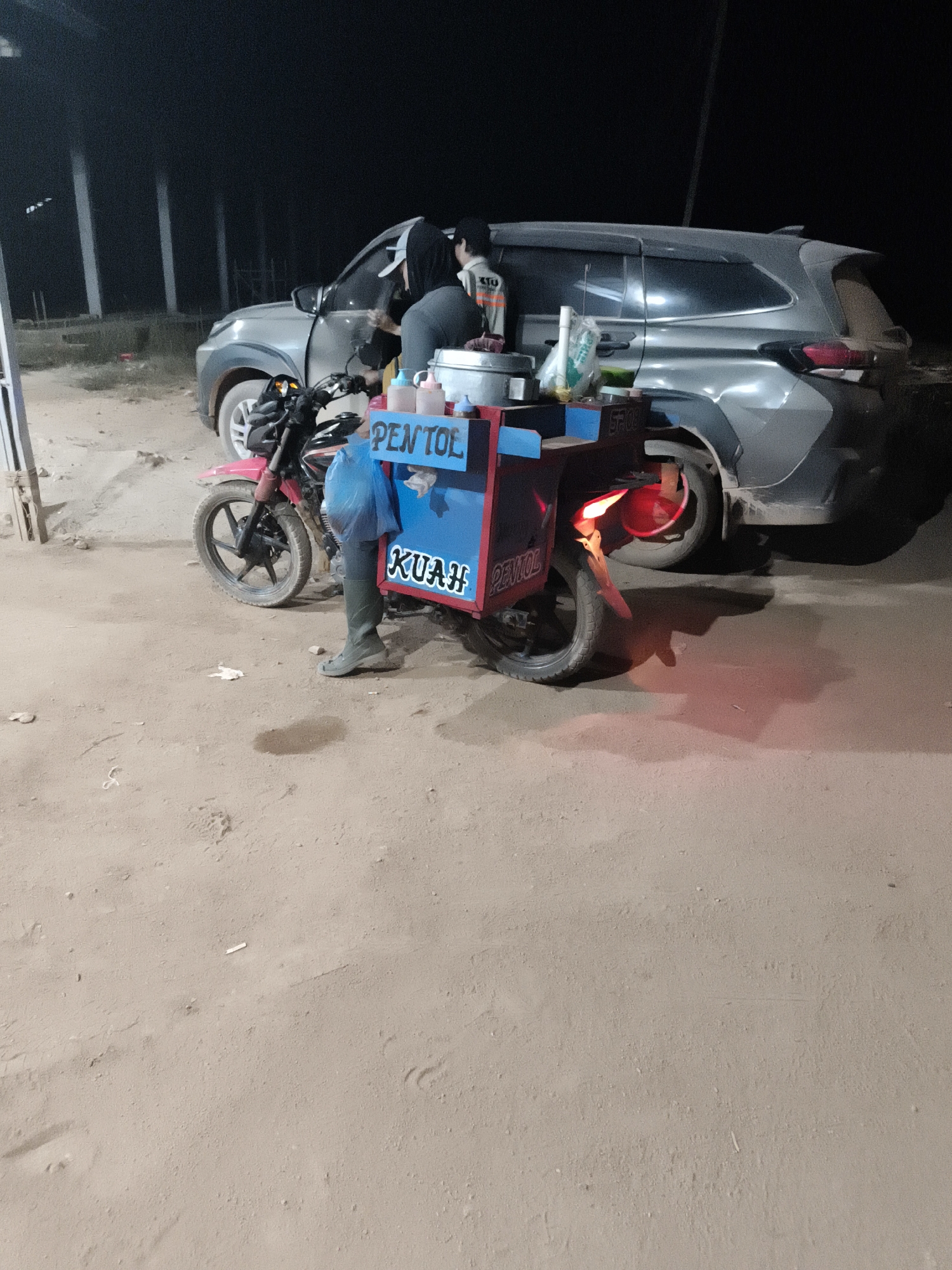 In the city of Selimantan Jaya, Indonesia, a street food vendor operates their business at night. A motorcycle, painted pink and black, is parked next to a gray SUV. Attached to the motorcycle is a food cart with a blue and red exterior. The cart is adorned with text that reads "PENTOL" in large blue letters and "KUAH" in smaller white letters. The vendor, wearing a dark hooded jacket and green boots, stands beside the cart. The ground is covered in sand, and the scene is illuminated by artificial lighting.