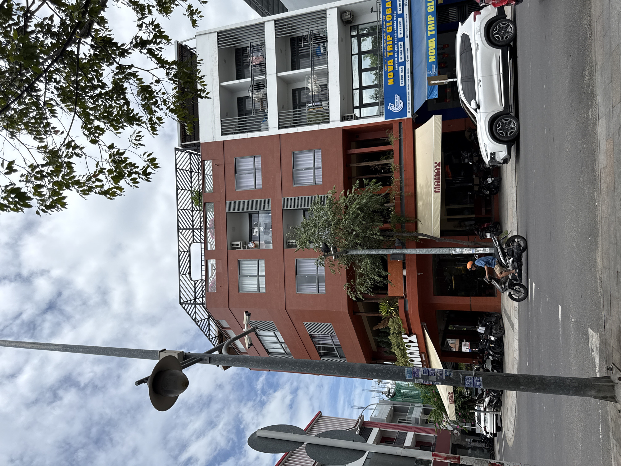 The image captures a street view in Nha Trang, Vietnam, featuring a modern multi-story building complex. The building on the left is a reddish-orange hue, with multiple windows and balconies. It houses a business named "MAMAX" with an awning over the entrance. Next to it, on the right, is a white building with balconies that appear to be offices for "NOVA TRIP GLOBAL," a travel company. The street in front is paved, with a sidewalk. A white SUV is parked on the right side of the street, and several motorcycles are parked in front of the MAMAX establishment. A person is riding a motorcycle down the street from right to left. The sky is cloudy. A lamppost with a security camera is positioned prominently in the foreground on the left side of the image.