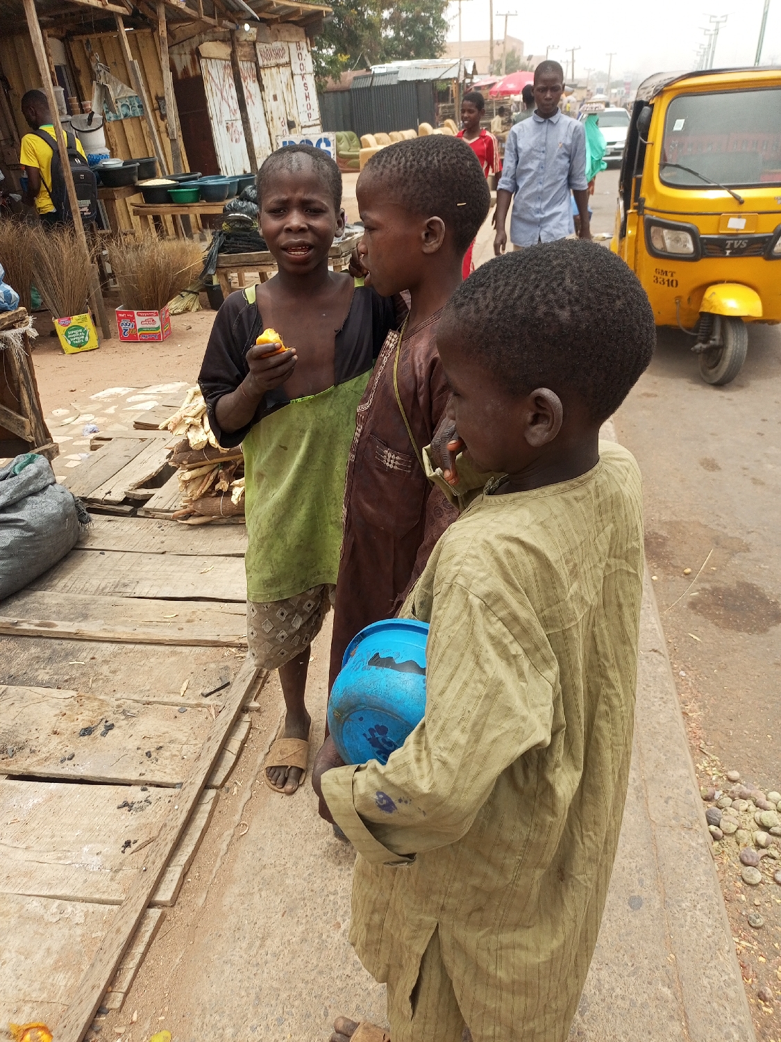 This candid photograph captures a street scene in Tempure, Nigeria, depicting three young boys in the foreground amidst a bustling market or roadside environment. The boy on the left, with dark skin and short black hair, wears a dark top with a light green apron-like garment over patterned shorts and sandals. He looks directly at the camera with an open-mouthed expression while holding a piece of orange fruit. Next to him, a boy in a brown long-sleeved tunic looks towards the third boy on the right. This third boy, mostly seen from behind, wears a light olive-green striped long-sleeved tunic and holds a blue plastic bowl.
In the background, a busy street scene unfolds with several adults and youths walking. A man in a yellow shirt stands near a wooden stall on the far left, while another man in a light blue shirt walks further into the background. A yellow auto-rickshaw, a common mode of transport, is parked on the right side of the street, displaying the text "TVS" and "GMT 5510".
The setting includes makeshift market stalls constructed from wood and corrugated iron, some appearing to sell various items like brooms and plastic bowls. One stall has "BOOTSHOP" written on it. The ground is a mix of dirt and raised wooden planks forming a pathway or display area. Cardboard boxes, including one for "MILO," are visible on the ground. The weather appears bright but hazy, typical of a dry, dusty climate, suggesting daytime. The scene conveys a sense of daily life and activity in a local market.