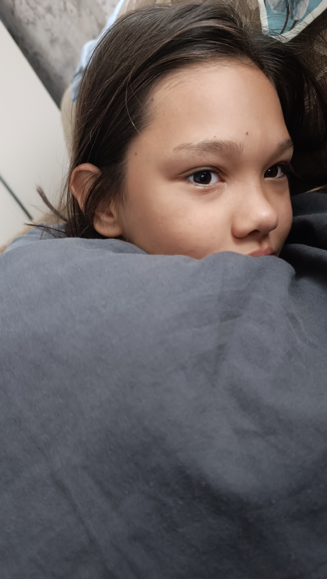 The image shows a close-up of a young person's face, partially obscured by a dark gray blanket or piece of fabric. The person has dark hair and brown eyes, and is looking off to the side. The background is blurred, but appears to be indoors. There is no discernible location in the city of Karsha, Russia.