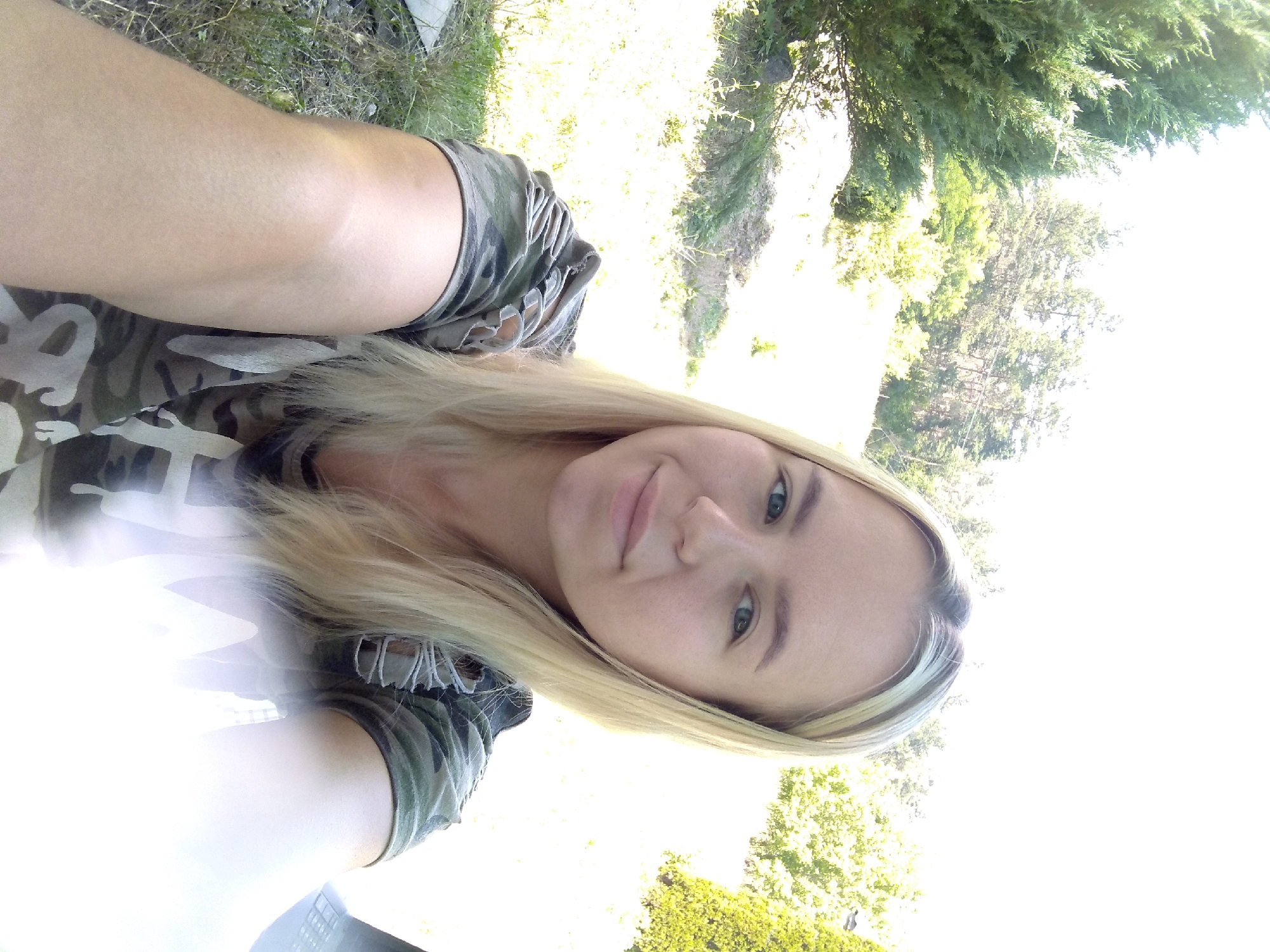 The image is a selfie taken outdoors, likely in or near Radisne, Ukraine. A young woman with blonde hair is the primary subject, positioned in the foreground and facing the camera with a slight smile. She is wearing a camouflage t-shirt with ripped sleeves. The background shows a natural environment with trees and grass, illuminated by bright sunlight. The overall impression is of a casual outdoor portrait, possibly taken during daytime in a rural or semi-rural setting.