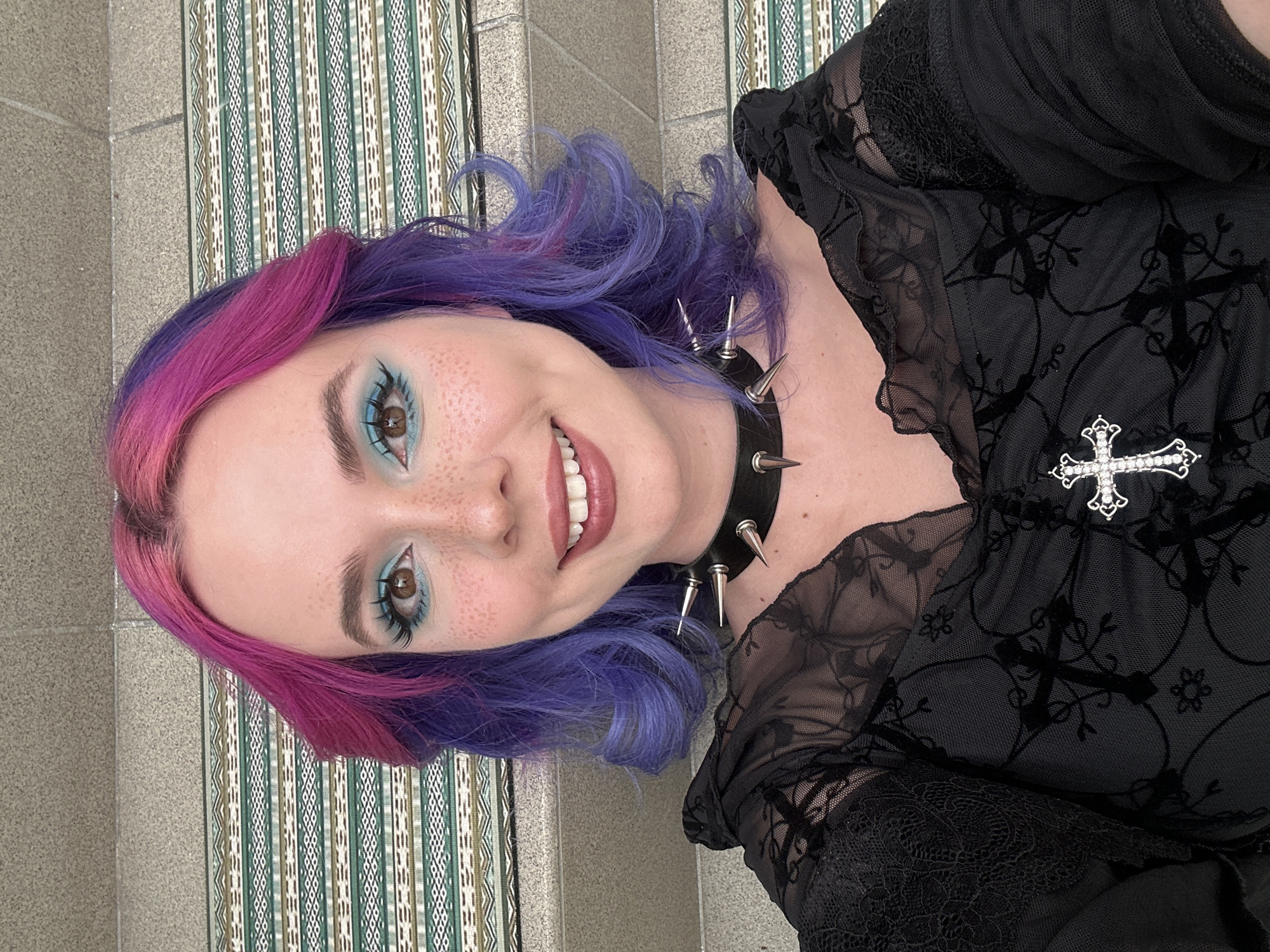A young woman is featured in a close-up portrait, smiling directly at the viewer. Her hair is dyed in a distinctive two-toned style, with bright pink color at the front and crown, transitioning to vibrant purple strands that frame her face and fall around her shoulders in soft waves. She has a fair complexion with noticeable freckles across her nose and cheeks. Her brown eyes are accentuated by bold turquoise-blue eyeshadow, dark winged eyeliner, and long, dark eyelashes. Her eyebrows are well-defined. A wide, genuine smile reveals her teeth.
She is dressed in a black top or dress that features sheer lace sleeves. The garment's main body is also black, adorned with a subtle pattern of crosses and possibly floral motifs. Around her neck, she wears a black choker embellished with several metallic spikes. A prominent, silver-toned cross brooch, intricately set with small clear jewels, is pinned to her top near her right shoulder.
The background includes a textured wall with grey-beige rectangular tiles on one side, and a section with a vertical striped pattern in shades of green, beige, and white on the other, resembling a decorative tapestry or patterned wall covering. The lighting is bright and even. The woman's attire and accessories suggest a gothic or alternative fashion style, contrasted by her cheerful expression. This scene is set in the City of Zagreb, Croatia. There is no visible text in the image.
