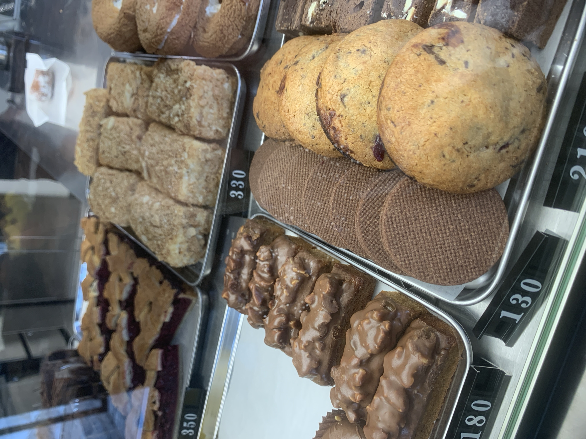 The image shows a display case in what appears to be a bakery or cafe. The focus is on various baked goods arranged in trays, including cookies, cakes, and bars. Price tags are visible near the trays, indicating the cost of each item. The location is assumed to be Izhevsk, Russia. There are no people visible in the frame.