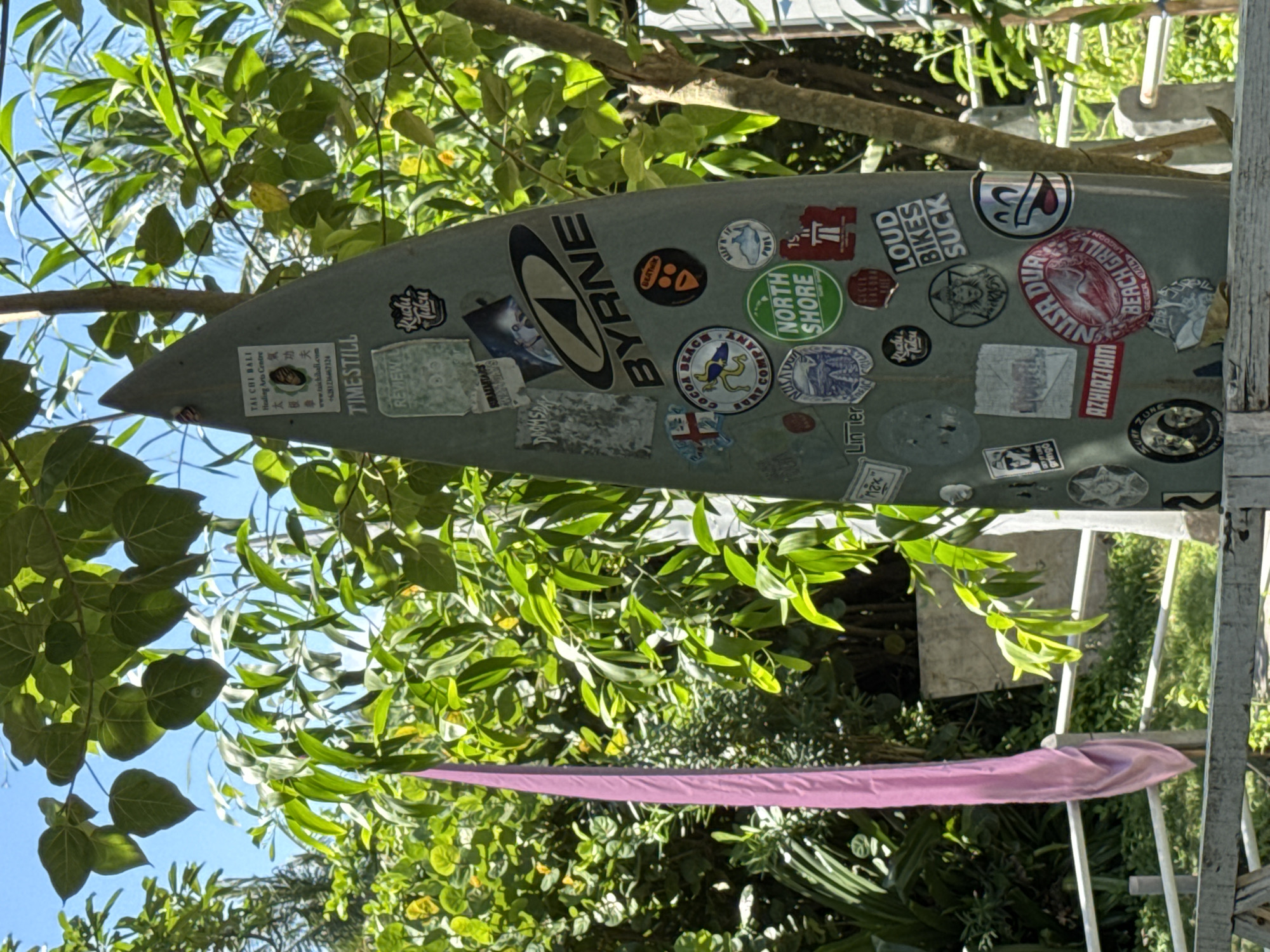 The image displays a sticker-adorned surfboard positioned vertically amidst lush green foliage under a clear blue sky. The surfboard, appearing light grey or green, is heavily decorated with numerous decals. Visible text from these stickers includes brand names and logos such as "BYRNE," "RUDU JUKU," "TIMESTILL," "DOOMSDAY," "BEACH GRILL," "ZUNE," and "SURAJASTARS."
Several stickers provide location cues, including "TAI CHI BALI Healing Arts Centre" with its website "www.taichibali.com" and phone number "+6281236467124." Other geographic references include "NORTH SHORE," "NUSA DUA," "AZHIAZIANS," and "GEGER 17T LAGOON." Phrases like "LOUD BIKES SUCK," "REVIEW," and "gringo" are also visible, along with others such as "BEATNIK COMPANY" and "LIN TER."
The setting is outdoors, likely in a tropical or subtropical region consistent with Kuta Selatan, Indonesia, indicated by the dense, vibrant green leaves and branches that partially frame the surfboard. The clear blue sky and bright illumination suggest it is daytime and sunny. A strip of pink fabric is visible towards the bottom right, hanging amongst the greenery. The image captures a static scene, showcasing a personalized surfboard in a natural, outdoor environment.