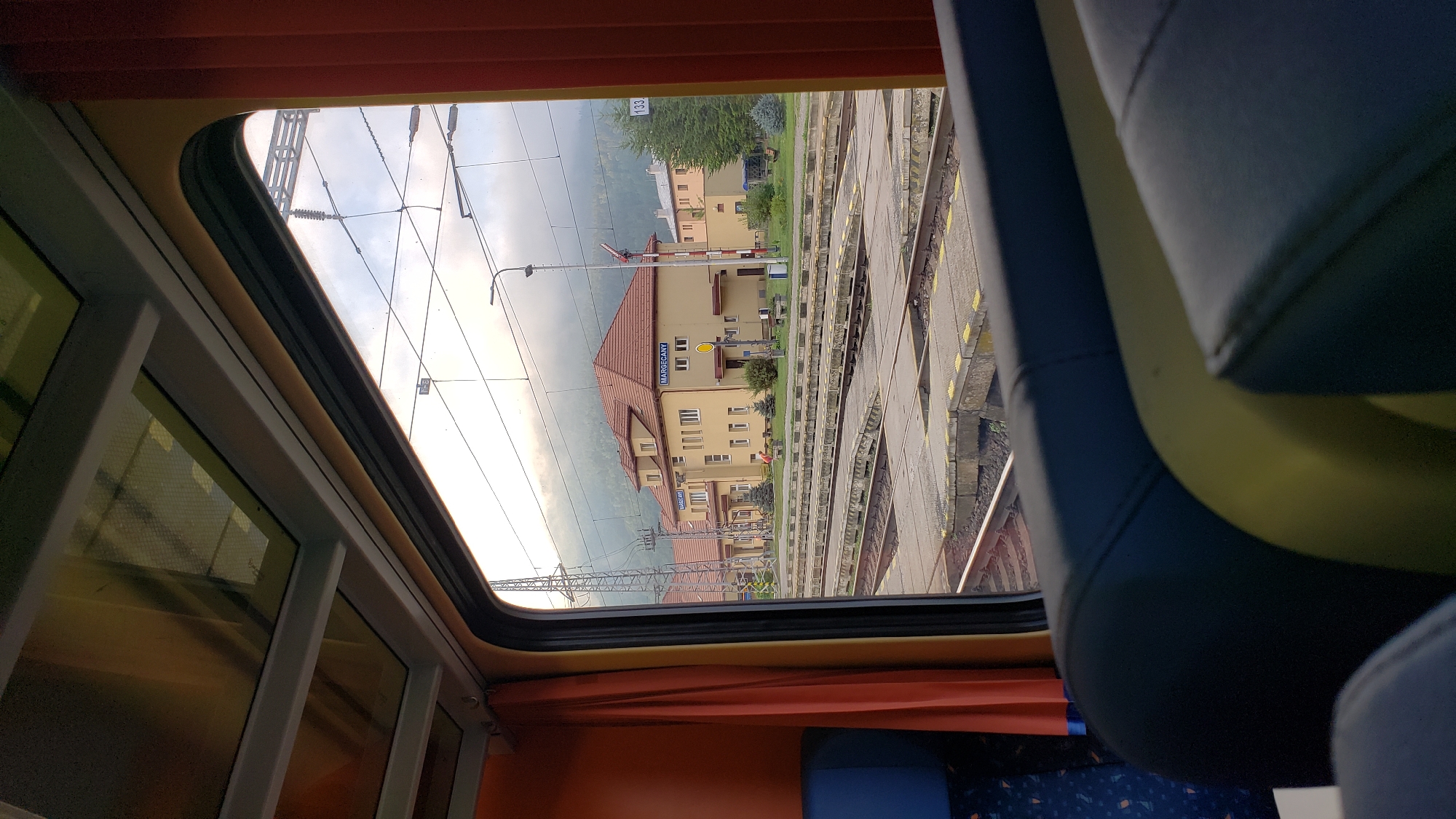 The image depicts a view from inside a train carriage, looking out at the Margecany train station in Slovakia. The foreground shows the interior of the train with dark blue seats and yellow accents. Beyond the window, the train station is visible, featuring a light yellow building with a red tiled roof. The building has multiple windows and signage indicating "MARGECANY". Train tracks and platforms are prominent in the mid-ground. Overhead, electrical wires and supports for the train lines are visible against a cloudy sky. In the background, a forested hillside rises under a hazy sky. Two individuals are visible on the station platform. One appears to be standing near a bench, and another is further away, possibly walking. The overall scene captures a moment at a rural Slovakian train station.