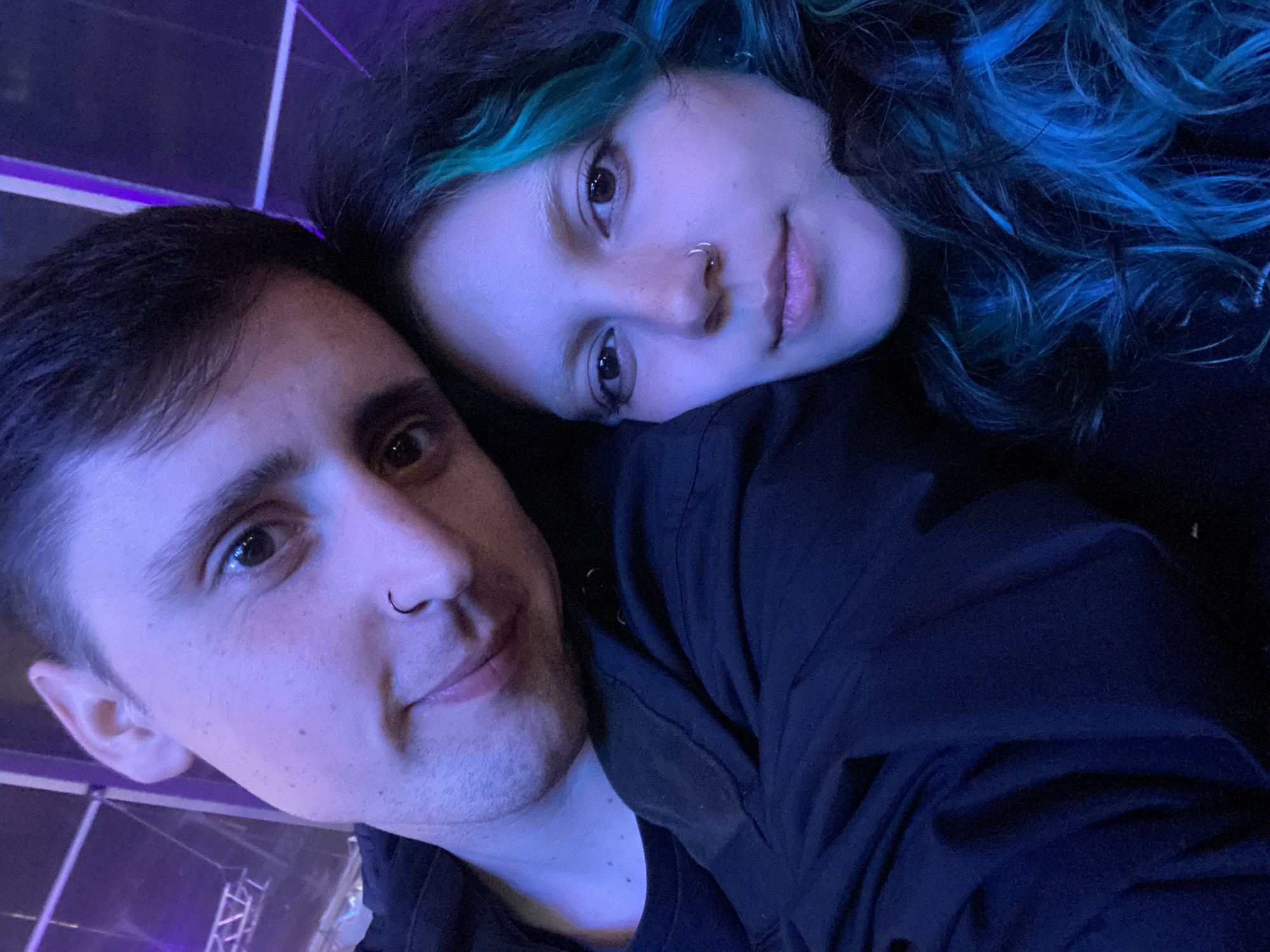 A couple poses for a selfie. The man is on the left with short dark hair, light skin, and a nose ring. The woman is on the right with multicolored dyed hair (blue and teal), light skin, and a nose ring. She rests her head on the man's shoulder. Both are wearing dark clothing. The lighting is dark with a blue/purple hue. The background appears to have stage lighting and rigging, suggesting a concert or performance venue, possibly located in Gijón/Xixón, Spain.