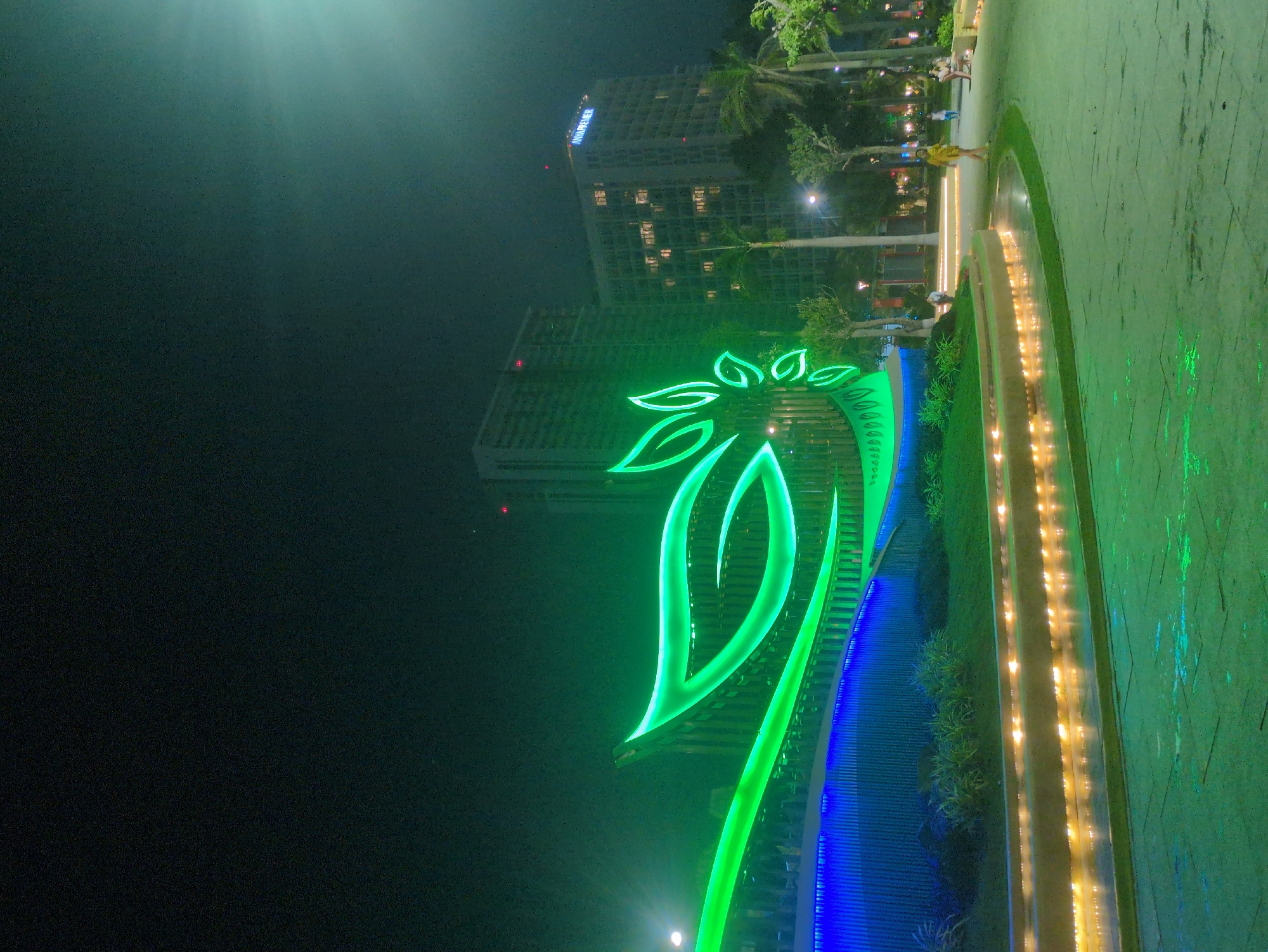 Captured at night in South Quy Nhon Ward, Vietnam, the image presents a brightly lit public space, likely a park or square. A stylized, luminous green palm tree sculpture stands prominently, framed against two tall buildings in the background, one labeled "MANDARIN." The ground is paved, with decorative illuminated strips and lush greenery interspersed. Several indistinct figures of people are observed in the background, engaged in casual movement, further suggesting a public gathering place. The artificial lights saturate the scene, creating a vibrant nocturnal urban landscape.