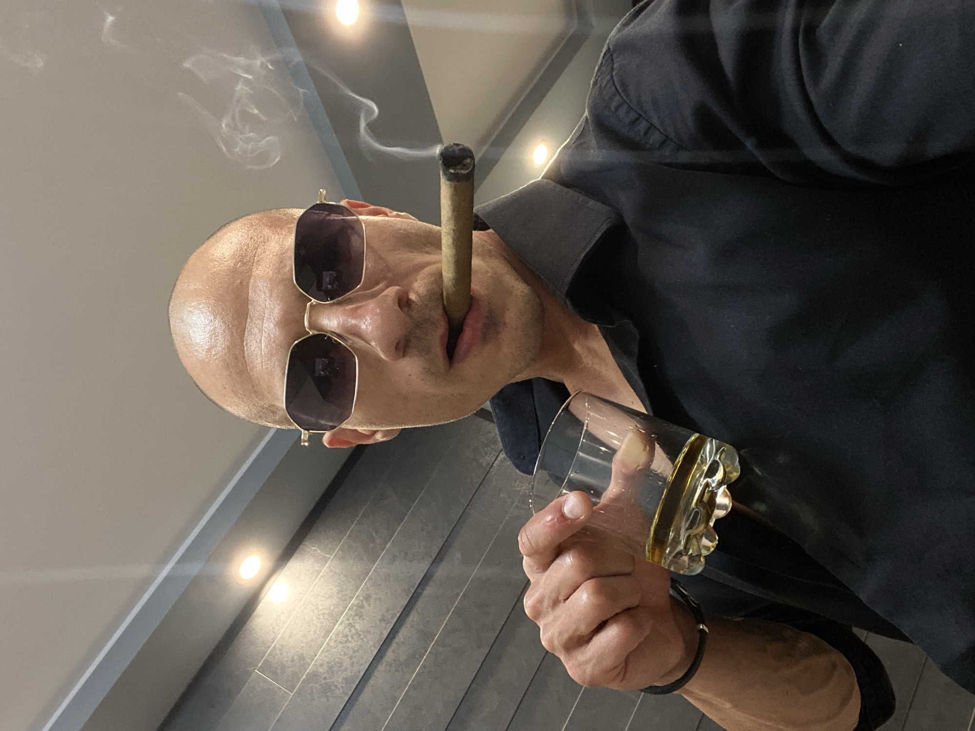 A bald man with sunglasses holds a glass of amber liquid in his left hand and a lit cigar in his mouth, smoke curling upwards. He is wearing a black collared shirt. The background consists of a textured wall and ceiling with lighting fixtures. The image is a close-up, self-portrait style shot. The specific location in the city of Pleven, Bulgaria cannot be determined from the visual information provided.