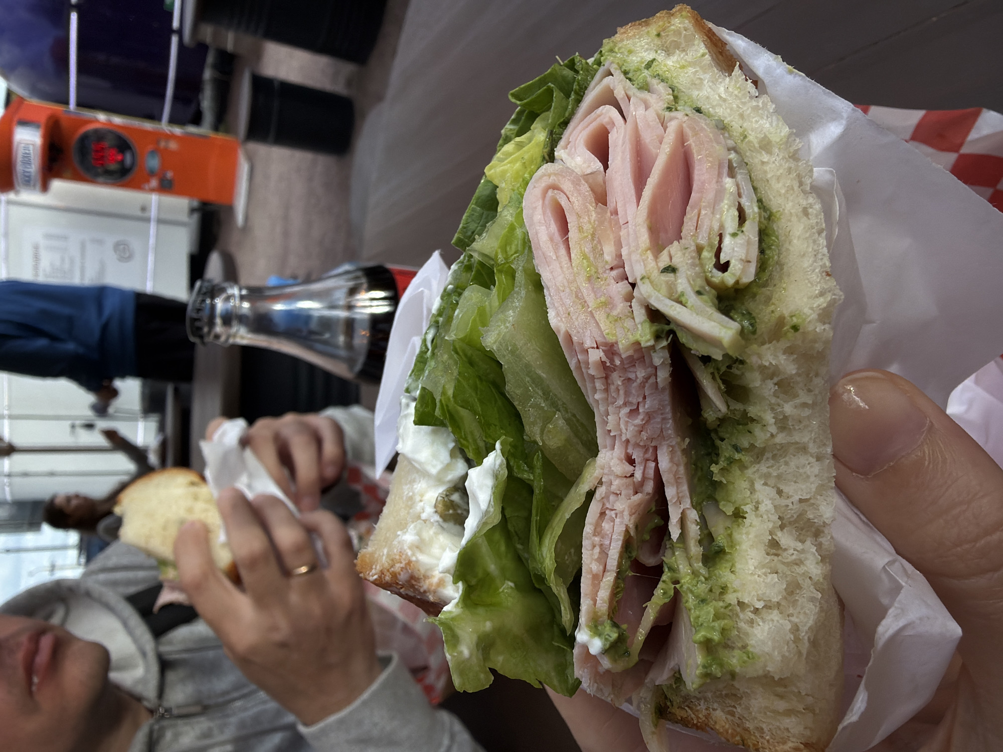 A person is holding a sandwich with layers of ham, lettuce, and a green spread, likely pesto, between two slices of bread. Another person is partially visible in the background, holding a similar sandwich. A glass bottle of Coca-Cola is present on the table. The setting appears to be a public space in Saint Petersburg, Russia, possibly a cafe or food court.