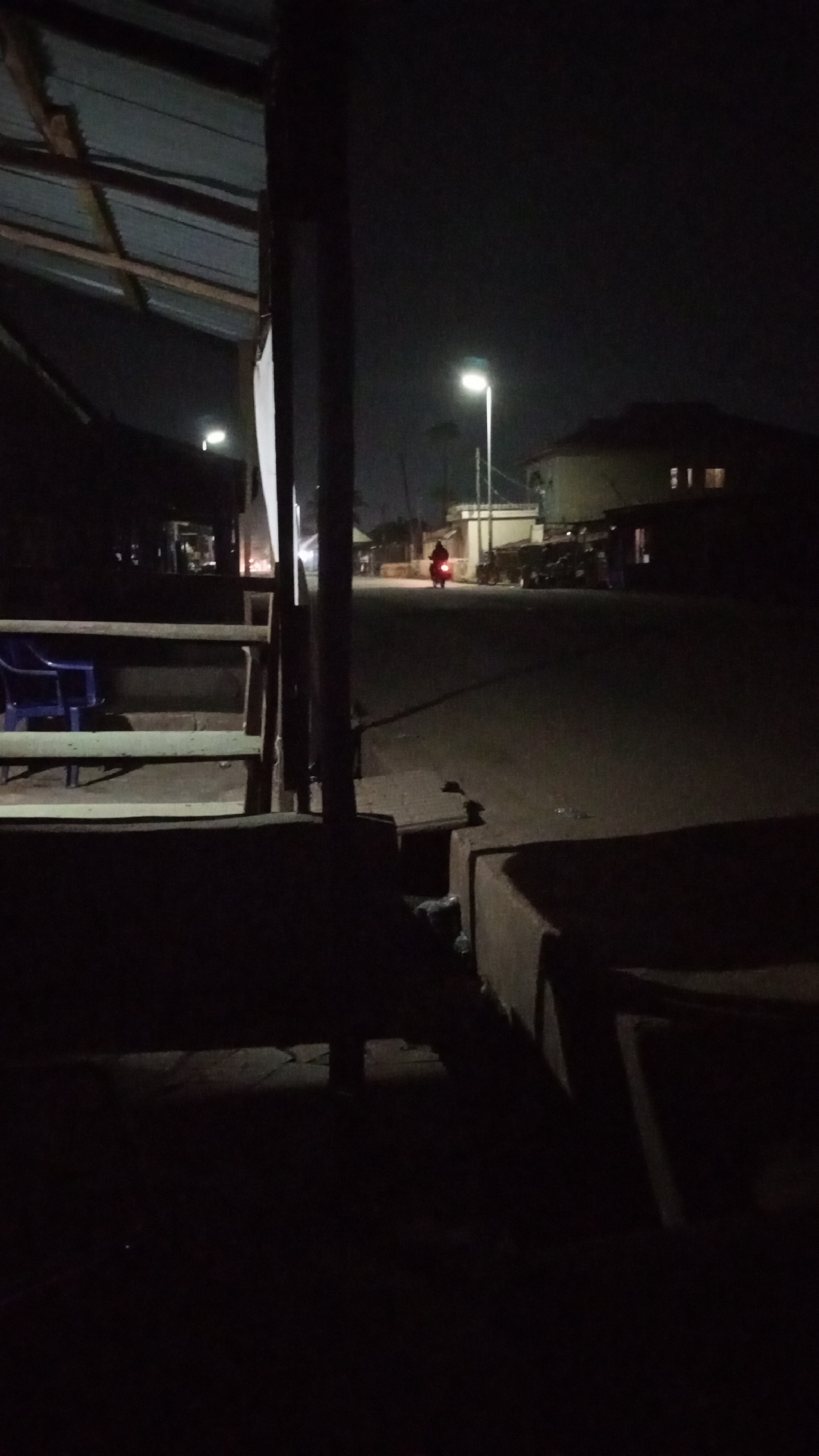 A nighttime scene in Sabon Tasha, Nigeria, viewed from a dimly lit, partially covered area. The foreground shows indistinct shapes of a structure, possibly a veranda or a shop front with wooden elements and a blue plastic chair. A road stretches into the distance, illuminated by intermittent streetlights. A person on a motorbike is visible further down the road. Buildings are silhouetted against the night sky on either side of the road.