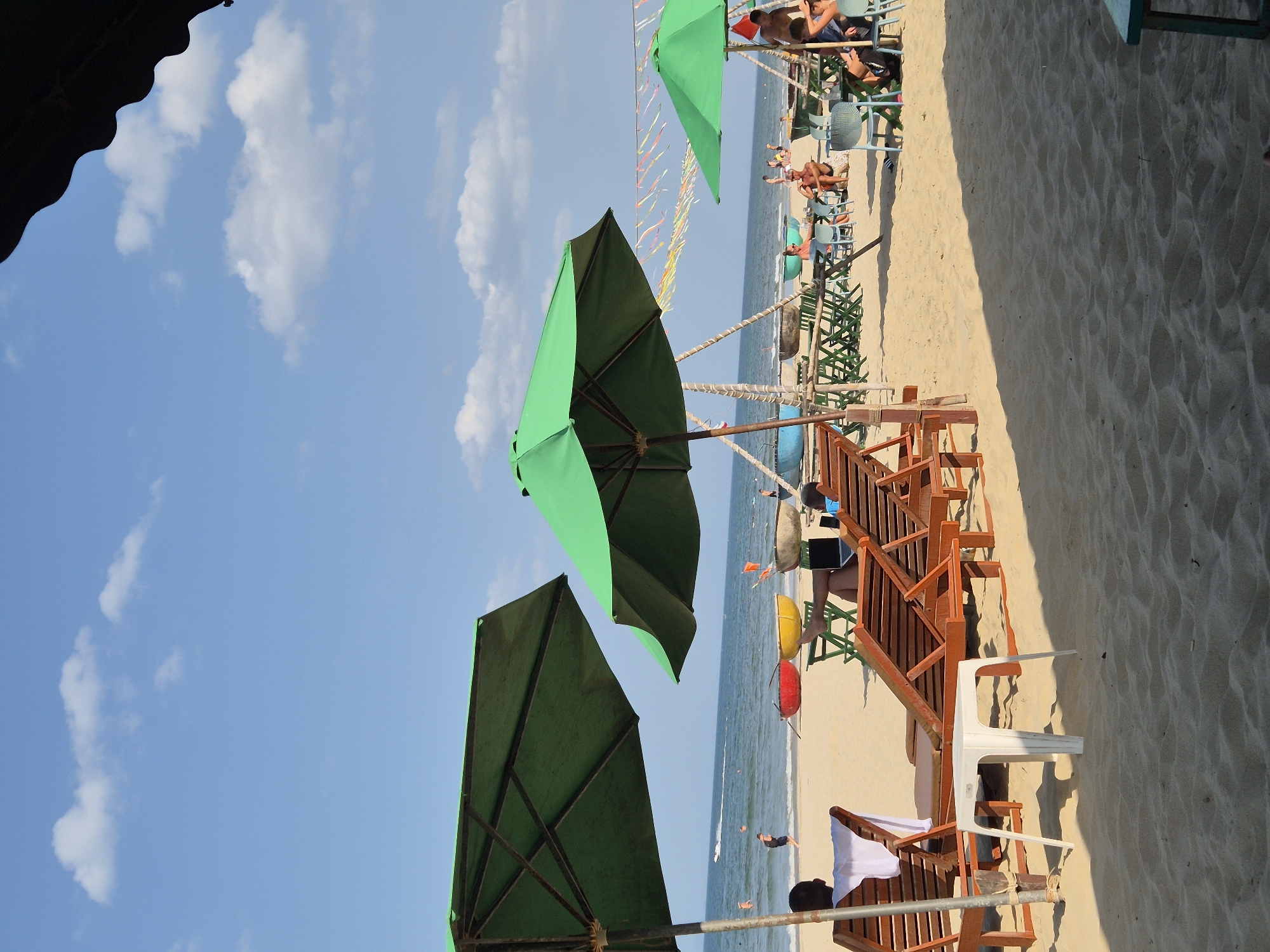 This is a photograph of a beach scene in Da Nang, Vietnam, on a sunny day with a clear blue sky and scattered white clouds. In the foreground, there are two large green beach umbrellas providing shade. The sand is a light golden color, and there are several empty wooden lounge chairs set up on the beach.
Beyond the lounge chairs, closer to the water, is a collection of tables and chairs, also set up on the sand. Some people are seated at these tables, while others are relaxing on the lounge chairs.
In the middle ground, the ocean is visible, with gentle waves rolling onto the shore. Several small, colorful boats are pulled up on the beach. People can be seen swimming and playing in the water in the distance.
In the background, there is a line of palm trees and what appears to be more beach infrastructure. Colorful streamers are strung between poles above some of the tables. The overall atmosphere is one of leisure and relaxation on a tropical beach.