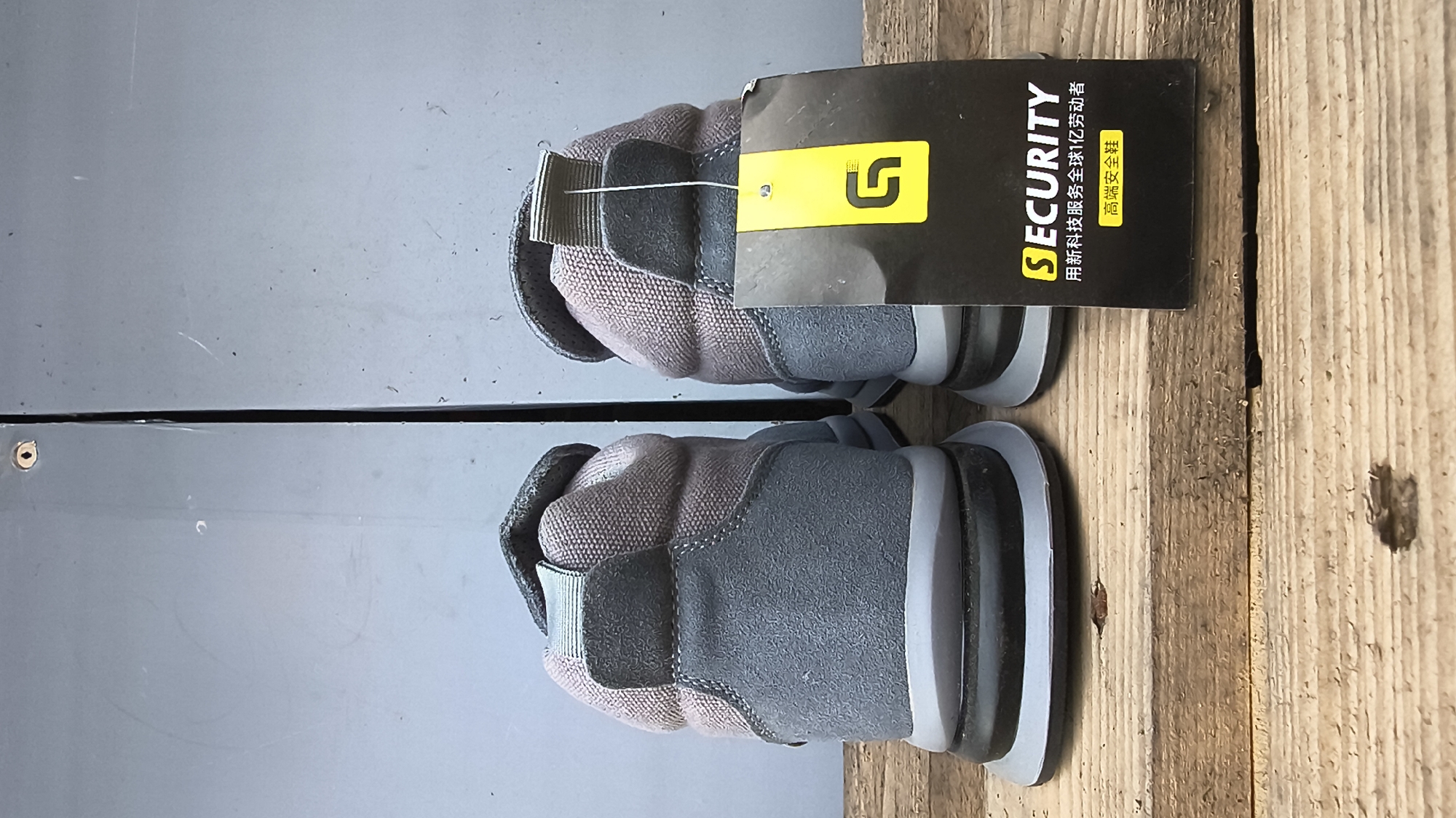 The image shows the back view of two grey and pink high-top sneakers, possibly safety shoes, standing on a weathered wooden surface. A black tag with a yellow "G" logo and the word "SECURITY" is attached to the right shoe. The tag also has Chinese text below. The background consists of a grey, possibly metal, surface with faint vertical lines. The scene is well-lit and appears to be an indoor or covered outdoor setting. There are no people visible in the image, and no specific landmarks or indications of Mikhaylovka, Russia, are present.
