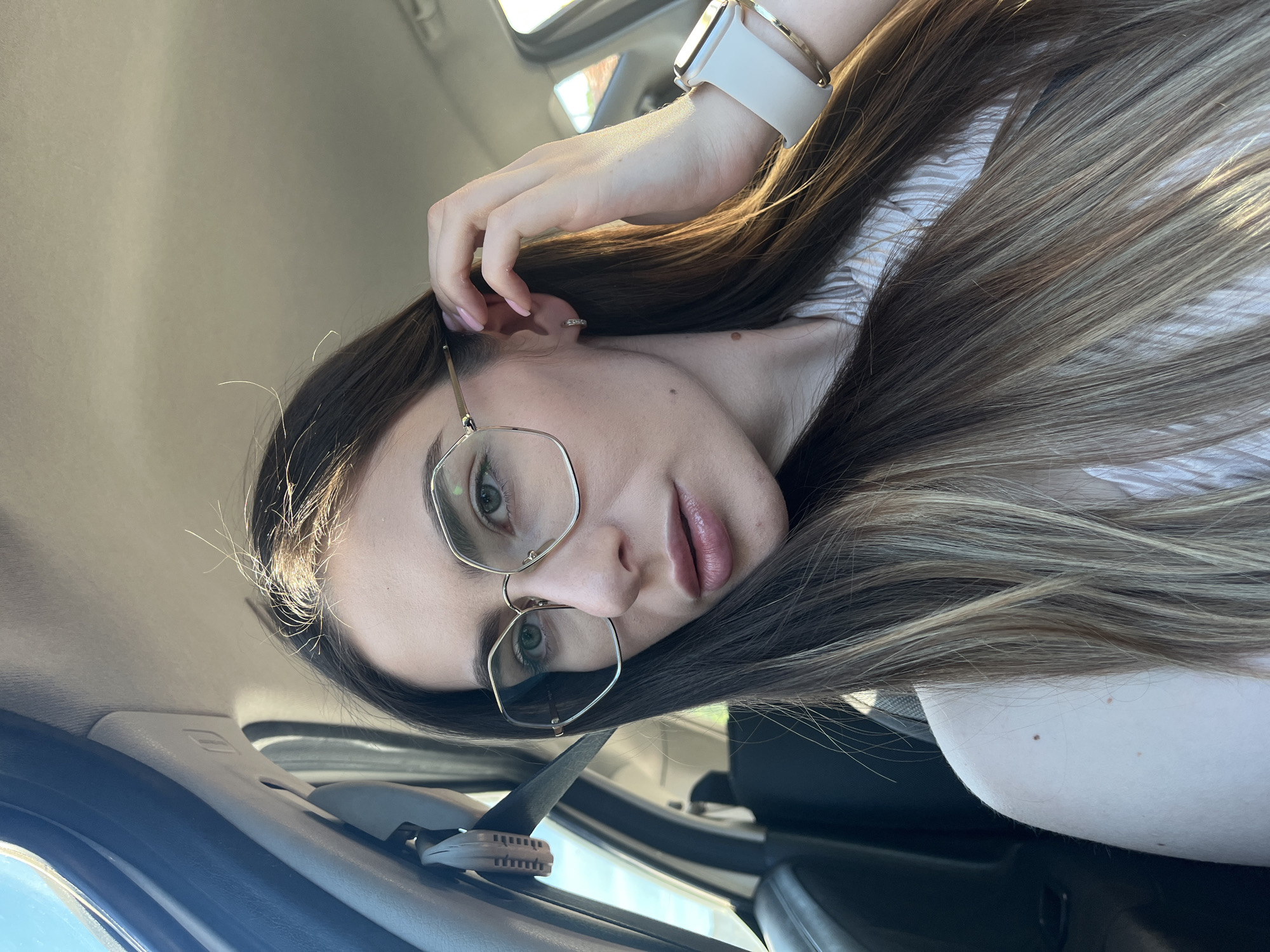 A young woman with long, brown hair and wearing glasses is in a car. She has a watch on her left wrist. The location is Unknown, Unknown. The subject is in the driver’s seat of a car, wearing a seatbelt. The interior of the vehicle is visible, including the dashboard and seats.