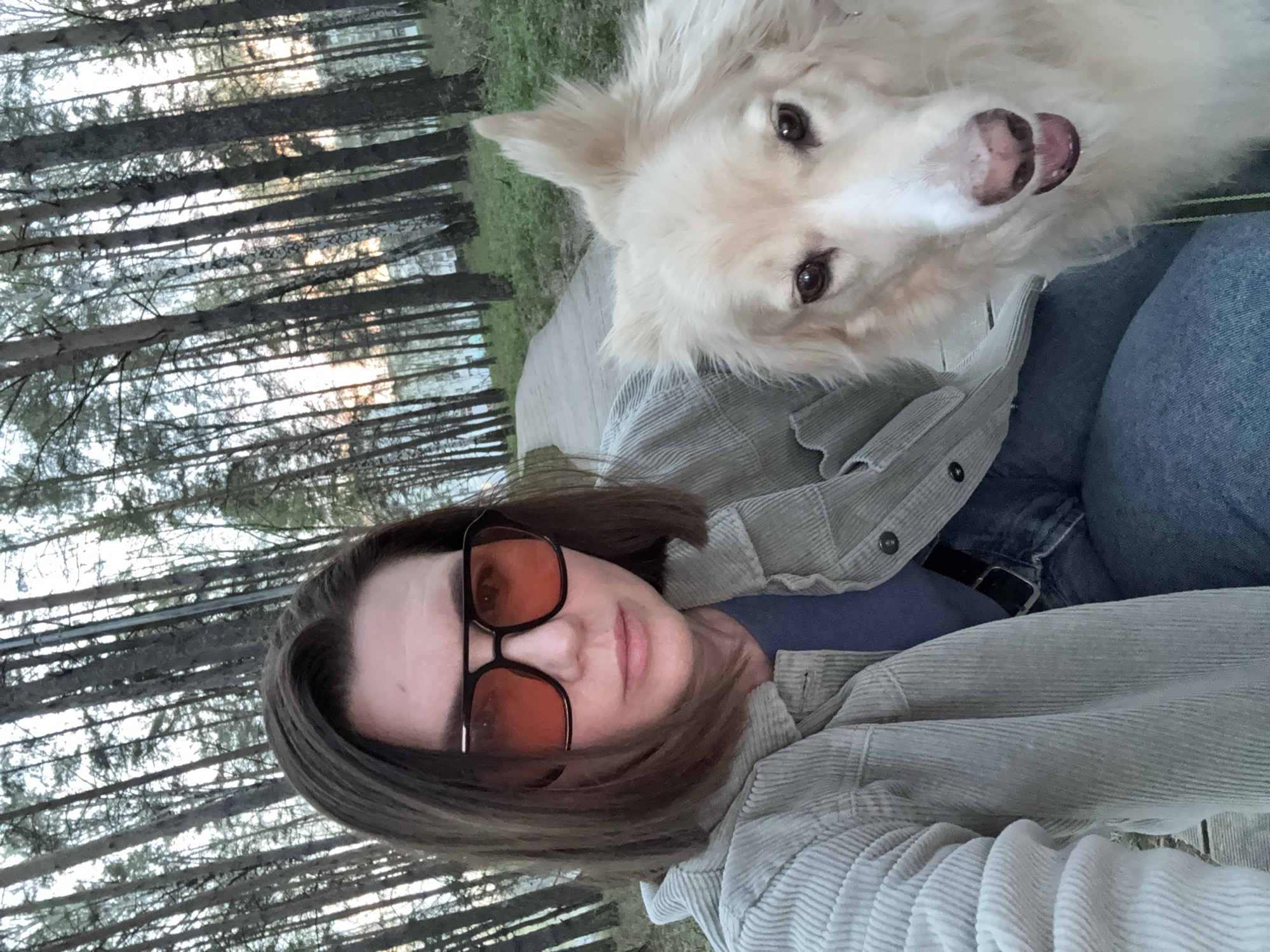 A woman wearing orange-tinted glasses and a light-colored corduroy jacket poses for a selfie with a white, fluffy dog. They are outdoors, and behind them is a sparse forest of slender trees, suggesting a park or wooded area. The woman is wearing a blue shirt and jeans. The photo may have been taken in a park or forested area within or near the city of Chaikovsky, Russia.