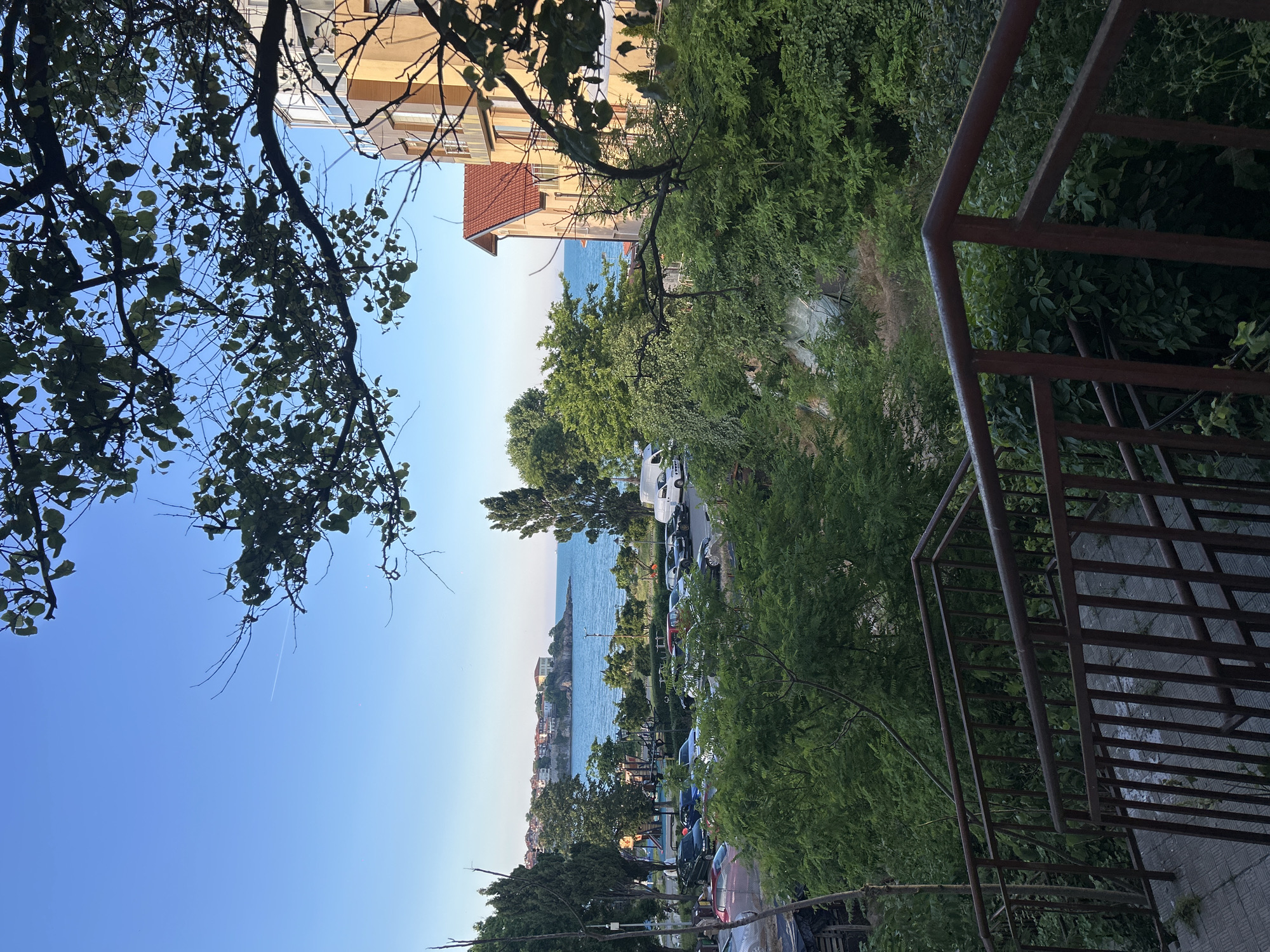 The image shows a view from a rusted metal staircase looking out over a street and the sea. The staircase is in the foreground, with a view of parked cars, trees, and buildings in the midground. In the background, the sea meets the horizon under a clear blue sky.
The location appears to be the city of Sozopol, Bulgaria, specifically an area near the coast with a view of the old town perched on a peninsula. The architecture of the buildings and the style of the cars suggest a Mediterranean or Eastern European coastal town. The presence of a playground on the left side of the midground indicates a residential or public area. The overall scene depicts a serene, late afternoon or early morning atmosphere. There are no people visible in the image.