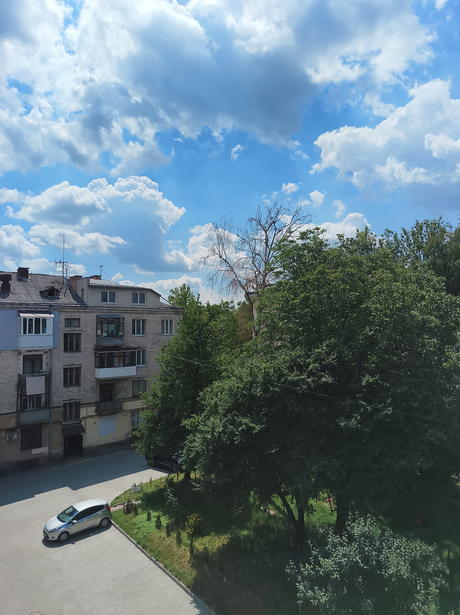 An aerial view captures a scene in Ternopil, Ukraine, dominated by a bright blue sky filled with fluffy white clouds. In the foreground, a paved courtyard area is partially occupied by a silver Ford Focus hatchback parked on the left side. Adjacent to the courtyard is a strip of green lawn with scattered shrubs and a small wooden structure.
Behind the lawn, a multi-story apartment building stands, constructed of light-colored bricks. The building features multiple windows and balconies. To the right of the apartment building, a dense canopy of lush green trees obscures parts of the structure and extends towards the right edge of the frame. A bare, spindly tree with light-colored branches stands taller than the surrounding foliage, positioned behind the apartment building and to the left of the dense trees. The overall scene suggests an urban residential area on a sunny day.