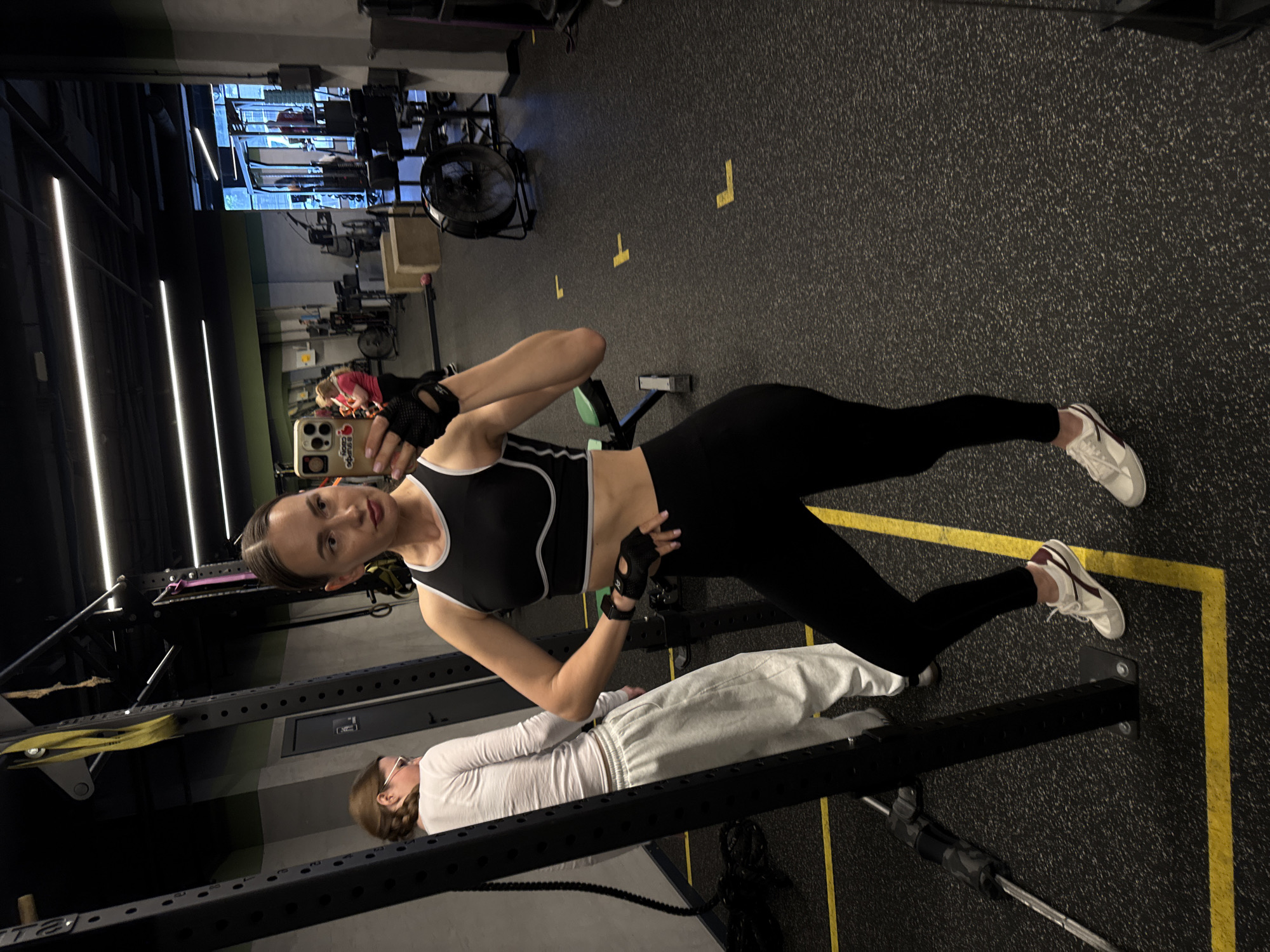 A young woman is captured in a mirror selfie inside a gym. She is the central subject, standing upright and looking directly at the camera (her reflection). She wears a black and white sports bra, black leggings, white sneakers with maroon accents, and black fingerless workout gloves, with a smartwatch visible on her left wrist. Her dark hair is styled back, and she has makeup on, including visible lipstick. She holds an iPhone with a light-colored case that has an indistinct graphic or text.
Behind and slightly below her, another individual is seen lying on a black workout bench, dressed in a light-colored long-sleeved shirt and light grey sweatpants, appearing to be resting.
The gym floor is a dark, speckled rubber material with bright yellow lines and 'L'-shaped markings. In the background, various workout equipment is visible, including power racks, a large fan, and other benches. The ceiling features long, bright linear LED lights, illuminating the indoor space. The image provides a glimpse into a workout session or a break within a gym environment in Sochi, Russia. No clearly legible text is present on any objects other than the indistinct phone case.