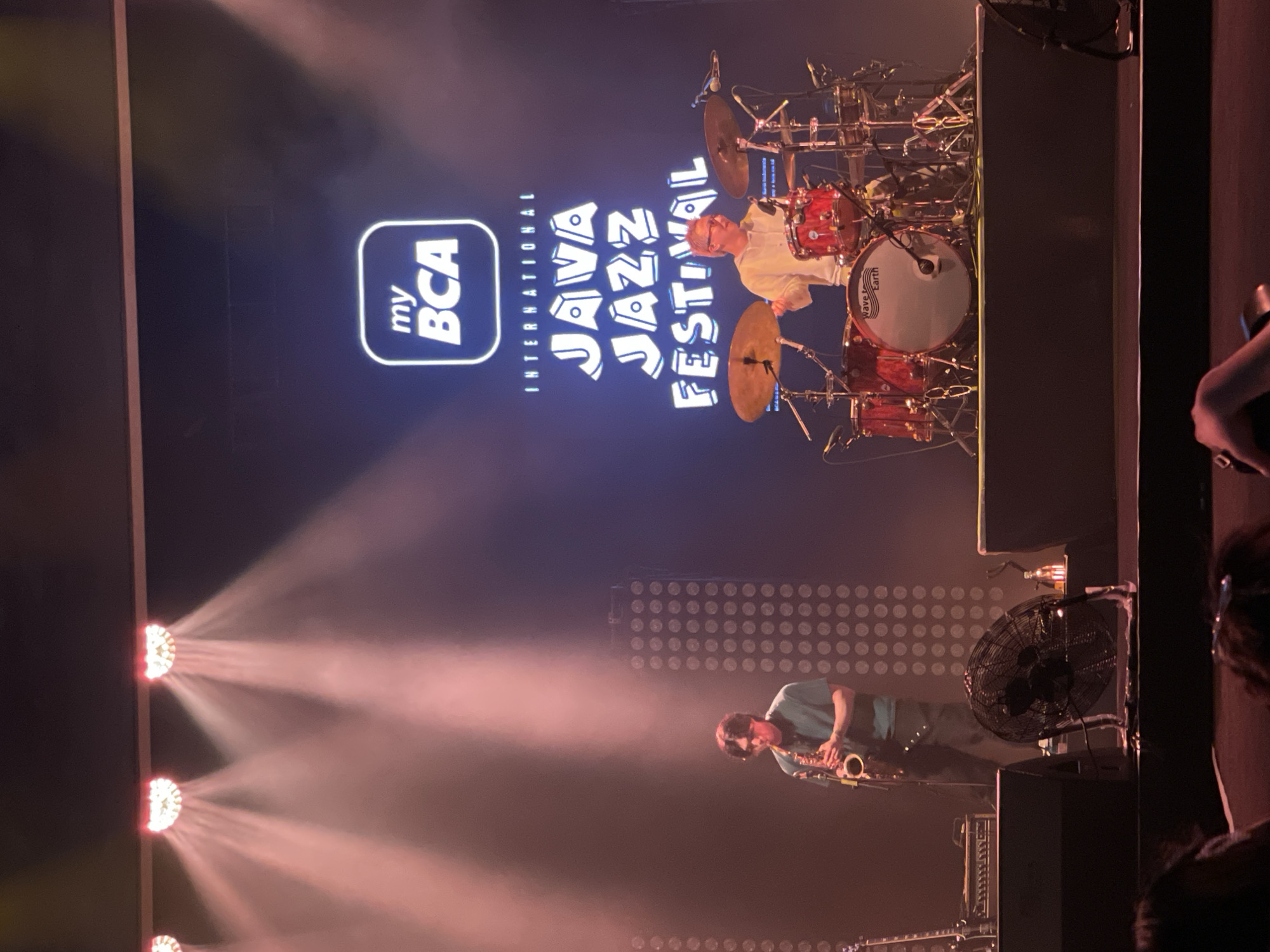 A music performance is underway, featuring a saxophonist and drummer, likely at the Salembaran International Java Jazz Festival in Indonesia, as suggested by the prominent "myBCA International Java Jazz Festival" logo projected on the screen behind the performers. The saxophonist, wearing a light blue t-shirt, is playing the saxophone, while the drummer is positioned behind a drum set. There are stage lights illuminating the scene, and a large fan is visible on stage. A portion of the audience is seen on the right side of the image.