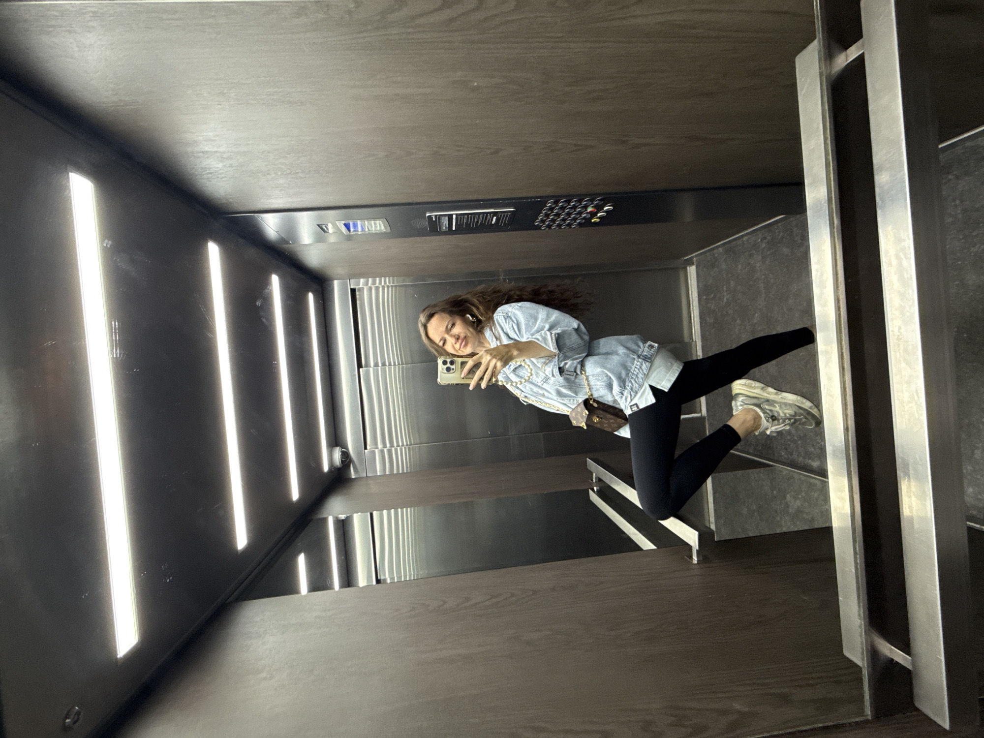 A woman is taking a mirror selfie in an elevator with wood-paneled walls and fluorescent lighting. She is wearing a light blue denim jacket over a white shirt, black leggings, and silver athletic shoes. She has a small brown crossbody bag with a gold chain. The elevator has stainless steel doors and handrails, and a control panel with buttons and a digital display. There is a security camera visible on the ceiling. The flooring appears to be a dark gray speckled material.