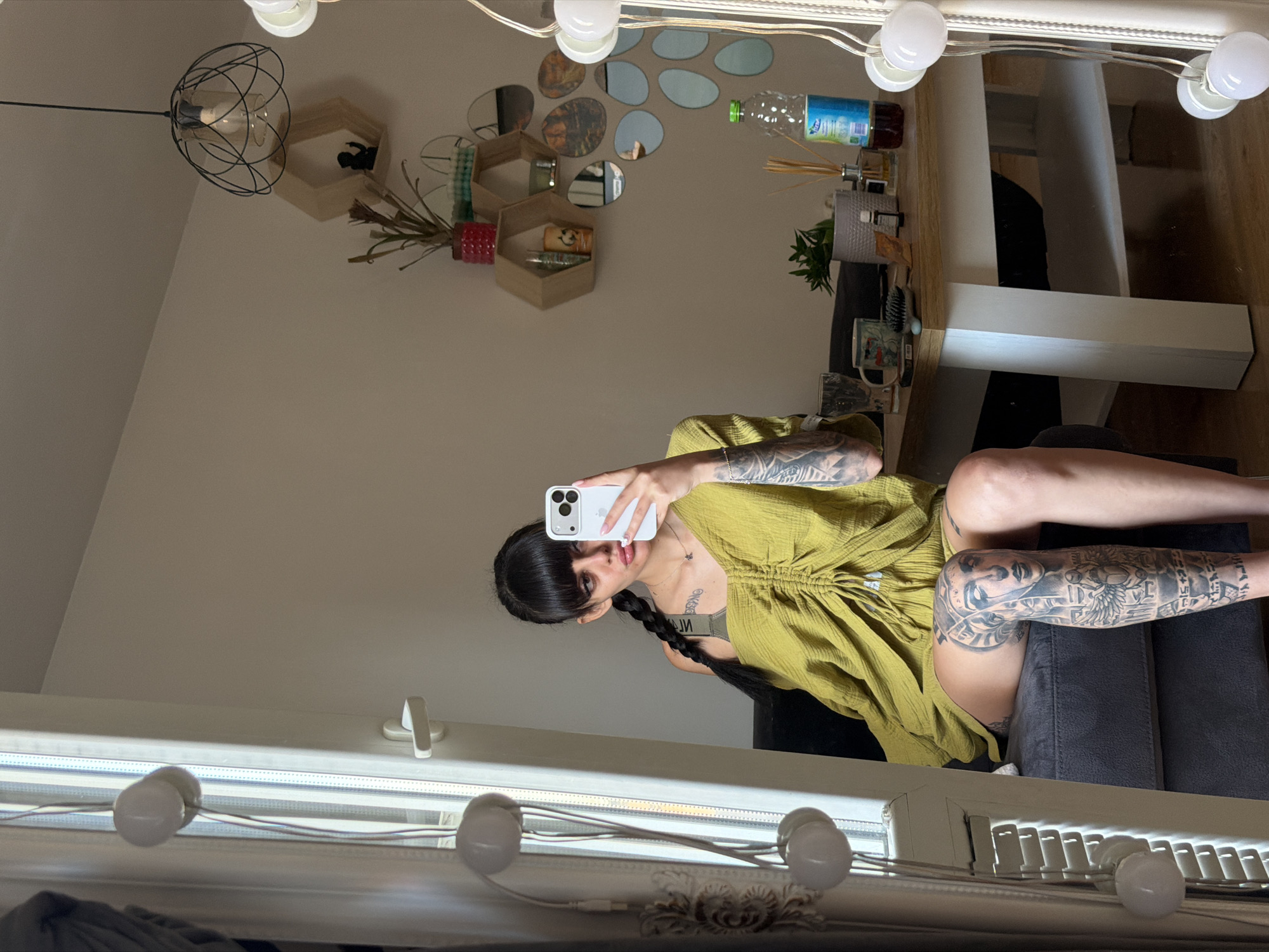 A young woman with dark hair styled in bangs and a braid is sitting on a grey couch, taking a selfie with her smartphone. She is wearing a lime green, ruched top and has extensive tattoos on her arms and legs, including a detailed portrait on her right leg. The woman is positioned in front of a window with vertical blinds and a string of white globe lights. To her right is a wooden dining table with a white base, on which sit a mug, a hairbrush, a bottle of drink, and a diffuser with reeds. Behind the table, on the wall, are hexagonal shelves displaying decorative items and small plants, and a collection of oval mirrors. The scene appears to be an indoor setting, likely a living room or dining area, in a city apartment. The image does not contain any information to place it in Plovdiv, Bulgaria.