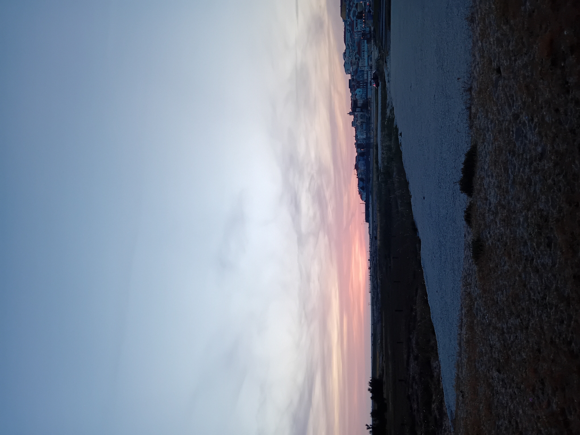 This media file captures a serene landscape scene at dusk or dawn. The sky, dominating the upper portion of the image, displays a beautiful gradient of colors, transitioning from a soft blue-grey at the top into vibrant pinks, oranges, and light purples near the horizon. Soft, wispy clouds are subtly streaked across these colorful hues.
Along the horizon, a distant city or town is visible as a dark silhouette against the glowing sky. Numerous buildings form a dense cluster, and faint lights twinkle within the city outline, suggesting habitation and the low light conditions of twilight.
Below the city silhouette, the landscape consists of distinct layers. A wide, lighter-colored strip, appearing to be a sandy beach, gravel bank, or dry riverbed, stretches horizontally across the middle ground. In the immediate foreground, a darker, textured area, likely dry grass, scrubland, or earth, occupies the lower part of the image. Some sparse, dark vegetation, possibly bushes or small trees, can be seen within this foreground.
There are no people or discernible activities taking place, and no visible text. The overall atmosphere is tranquil and peaceful, characterized by the soft, muted lighting and the beautiful sky colors. Given the context of Figueira da Foz, Portugal, this scene likely depicts a view across the Mondego River estuary or a coastal area at sunset or sunrise.