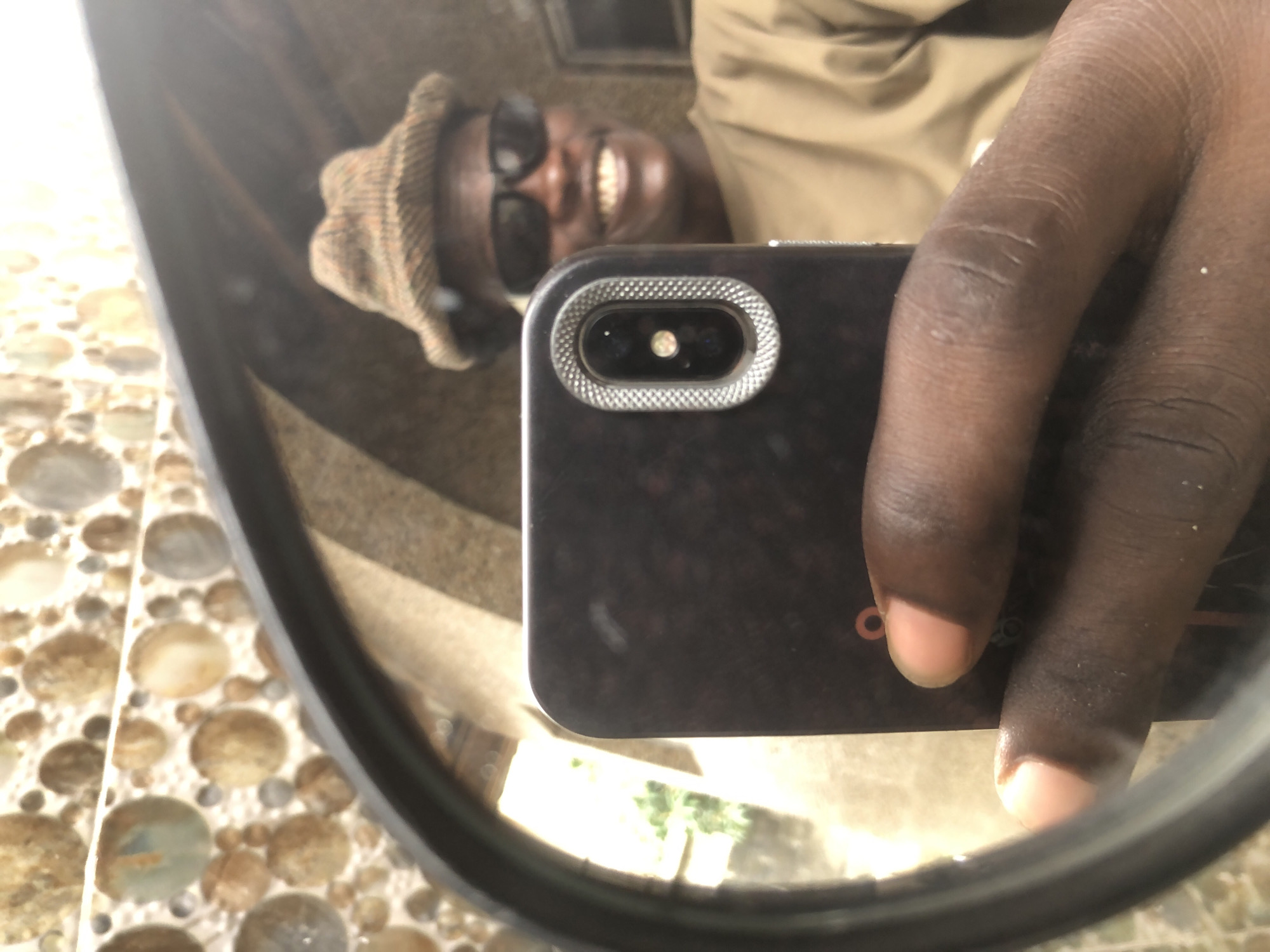 In Ajah, Nigeria, a person is captured in a mirror selfie. The individual, wearing a brown plaid hat and dark sunglasses, is smiling. They are holding a smartphone, pointed towards the mirror to take the photo. The background shows a wall with circular textured tiles and an arched opening. The overall lighting suggests an indoor setting, possibly a bathroom or a room with reflective surfaces.