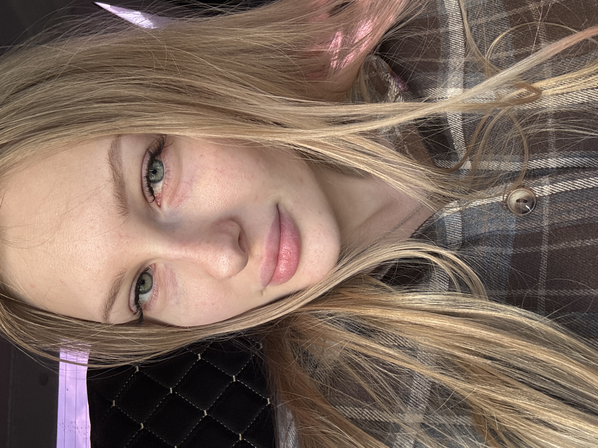 A selfie of a young woman with light blonde hair is shown, presumably taken indoors. Her hair is long and styled loosely, with some strands falling across her face and chest. The woman has fair skin, green eyes, and slightly pink lips. She is wearing a brown and gray plaid shirt with a button visible on the right side of the frame. The background features a quilted black surface, possibly a car seat or other upholstered item. The location of Perm, Russia is not discernible.