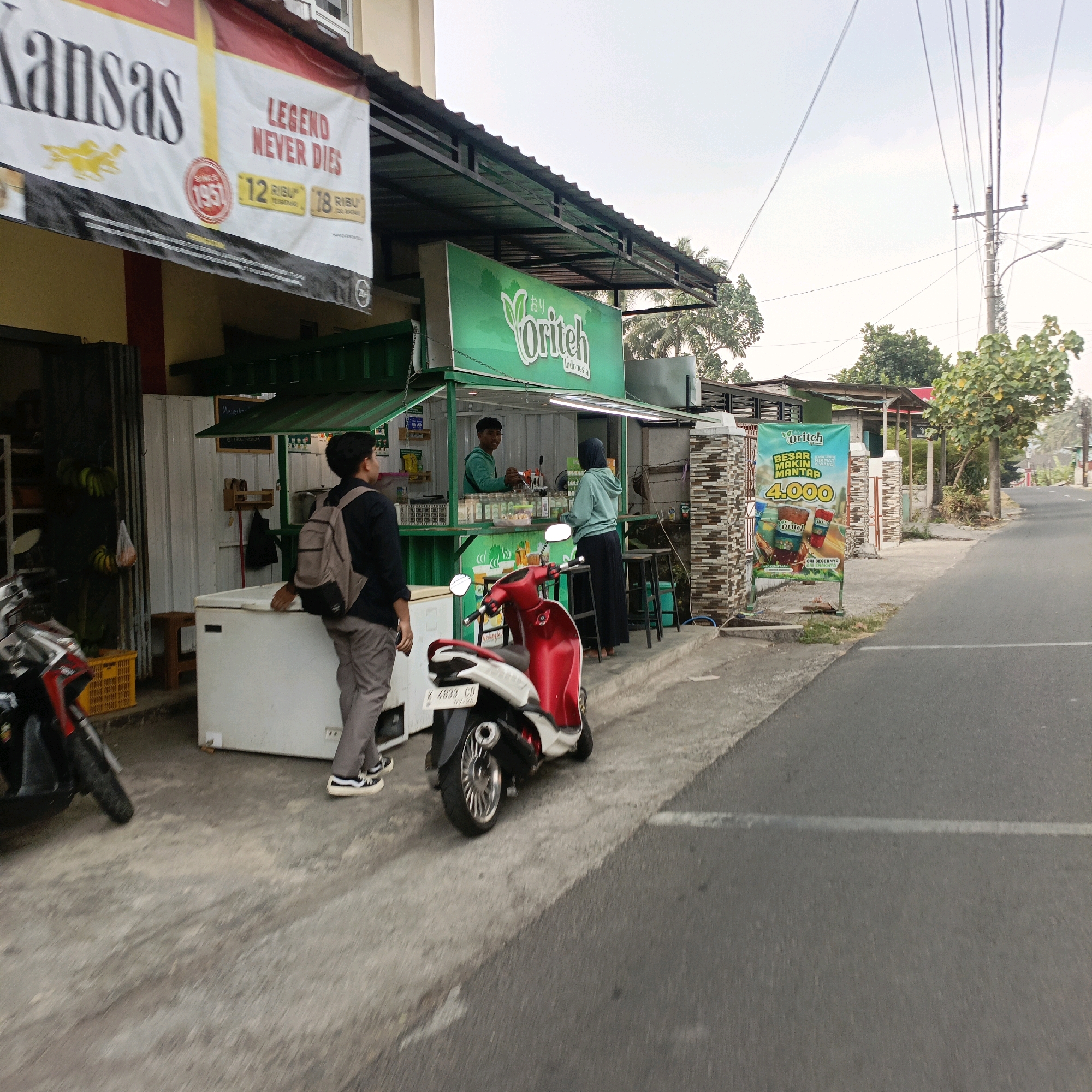 In Salatiga, Indonesia, a street-level view captures a small roadside beverage stall, prominently displaying a green sign that reads "Oriteh Indonesia." The stall is open, with a vendor and a customer interacting. In front of the stall, a red and white scooter is parked. To the left, a man with a backpack stands near a large white freezer. The background shows a street, utility poles with overhead wires, and trees. A large banner advertising "Kansas" and "Legend Never Dies" hangs on the building to the left of the stall. Another large banner advertising "Oriteh" with pricing is displayed on the right side of the scene. The environment suggests a warm, possibly humid, day.