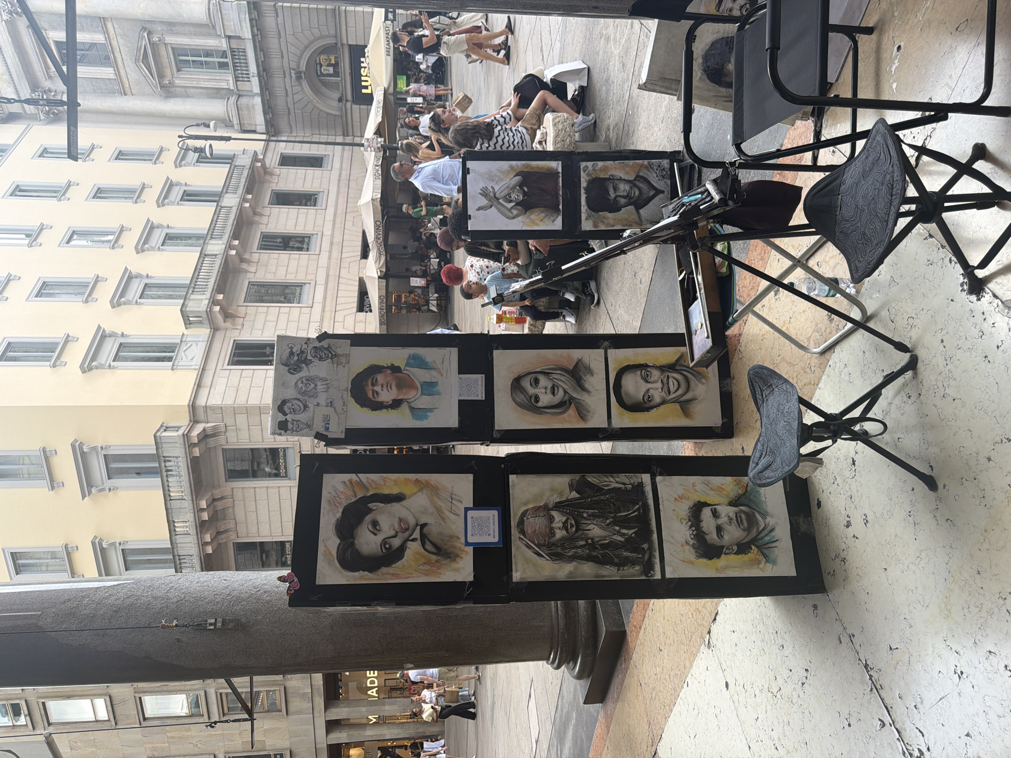 The image depicts an outdoor scene on a stone-paved street in Milan, Italy, likely in a pedestrianized area given the presence of shops and cafes. In the foreground, an artist's stall is set up with several framed portraits displayed vertically on black boards. The portraits are rendered in charcoal or graphite and depict famous personalities, including what appears to be Angelina Jolie, Johnny Depp as Captain Jack Sparrow, and Diego Maradona. To the right of the artwork display, a portable easel holds painting supplies, and two foldable chairs are set up for seating.
In the mid-ground, a variety of people are present, some walking along the street, while others are seated at outdoor tables belonging to a cafe with umbrellas. The architecture in the background features buildings with multiple stories, adorned with decorative balconies and large windows, characteristic of Milanese urban design. The overall atmosphere suggests a bustling public space where art and commerce intersect.