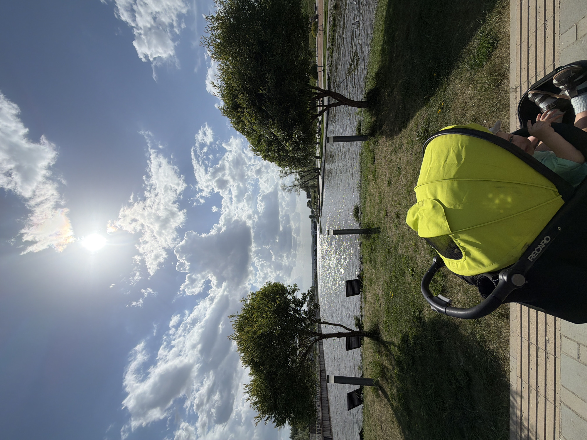 A sunny outdoor scene depicts a baby in a lime-green Recaro stroller positioned on a brick walkway, likely in Votkinsk, Russia. The stroller is partially obscured. In the background, a body of water, potentially a river or lake, glistens with sunlight. Two trees are visible on the shore, casting shadows onto the grass. Benches and light posts line the water's edge. The sky is blue with scattered white clouds, and the sun shines brightly.