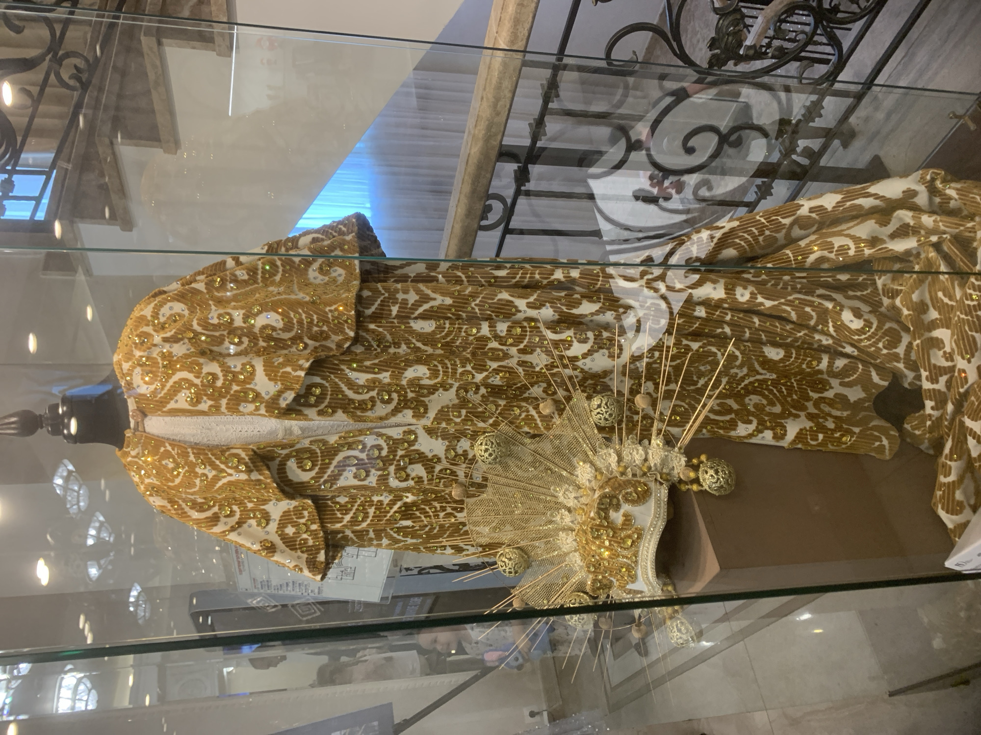 The image shows a gold and white ornate costume displayed within a glass case. The costume includes a jacket, dress, and a golden headpiece with radiating rods and decorative balls. The display is located inside a building, and a portion of an ornate metal railing is visible in the background. Given the image source, it can be inferred that the display is possibly located in Izhevsk, Russia.