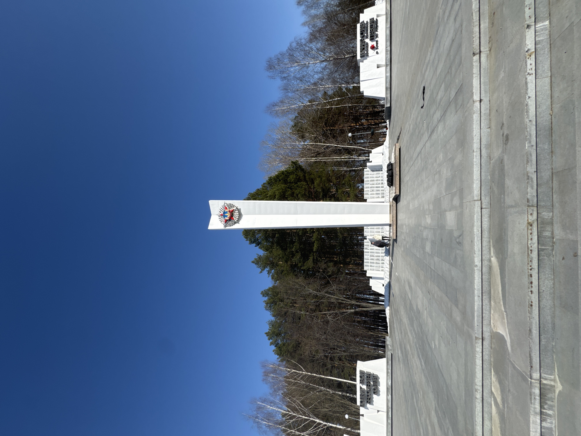 A clear, daytime view of the Chaikovsky Victory Memorial in Chaikovsky, Russia. The memorial includes a tall, white pillar surmounted by a coat of arms. A forest of evergreen trees forms the backdrop behind the memorial, and the surrounding area is paved with gray stones. No people are visible in the immediate scene. The sky is a clear, bright blue.