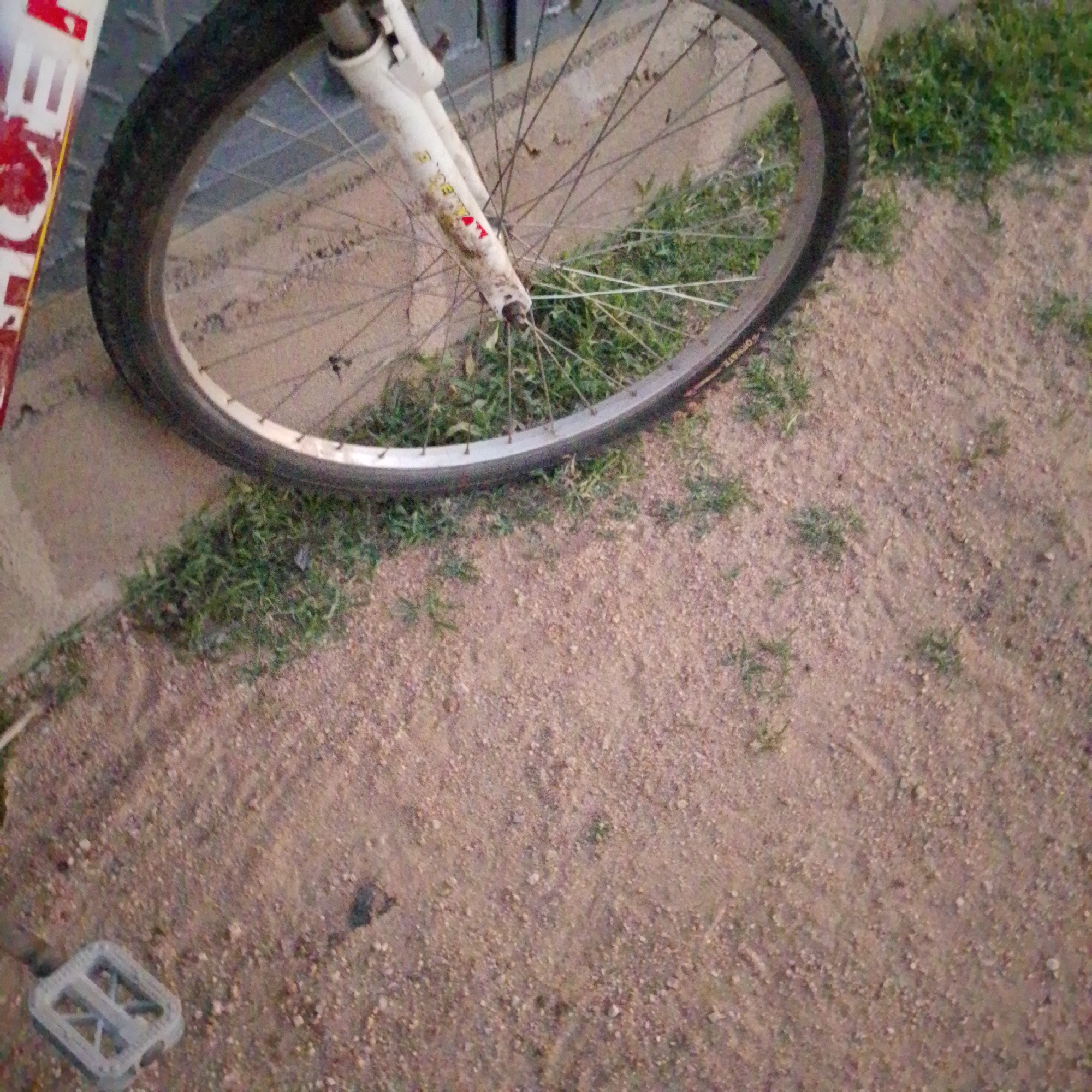 A close-up shot captures the front wheel and fork of a bicycle resting on a dirt and grass surface. The bicycle's wheel is a dark tire with a silver rim and numerous spokes. The front fork is white and appears dirty, with some red and yellow lettering visible. To the left of the wheel, a portion of the bicycle's frame, painted red and white, is partially visible. In the lower left corner of the image, a gray bicycle pedal is visible. The ground is a mixture of fine dirt and patches of green grass. The background consists of a textured, light-colored wall. The overall scene suggests an outdoor setting, possibly in a park or a yard. The image does not contain any people. The location is Wuro Allahamdu, Nigeria.