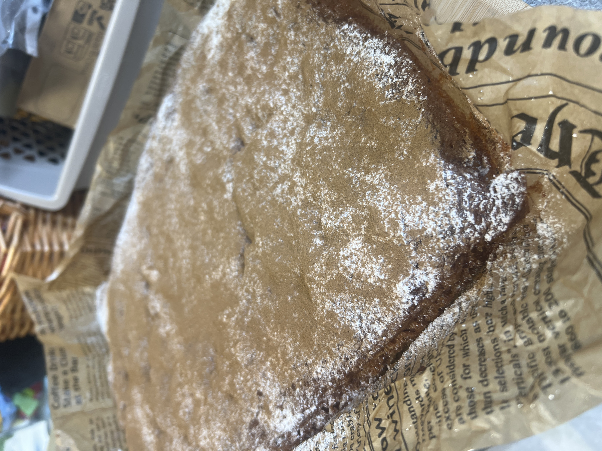 The image shows a rectangular cake dusted with powdered sugar and cinnamon, placed on a sheet of brown paper with text printed on it. The paper appears to be newspaper or wrapping paper. In the background, a wicker basket and a light-colored plastic container are visible. The scene is indoors, with diffused lighting. There are no people or animals present in the image. The cake is the primary subject, positioned centrally and taking up a significant portion of the frame. The focus is on the texture of the cake and the dusting of sugar and cinnamon. The environment is domestic or a casual food setting, possibly a kitchen or a cafe. No information is available to determine the location in Khabarovsk, Russia.