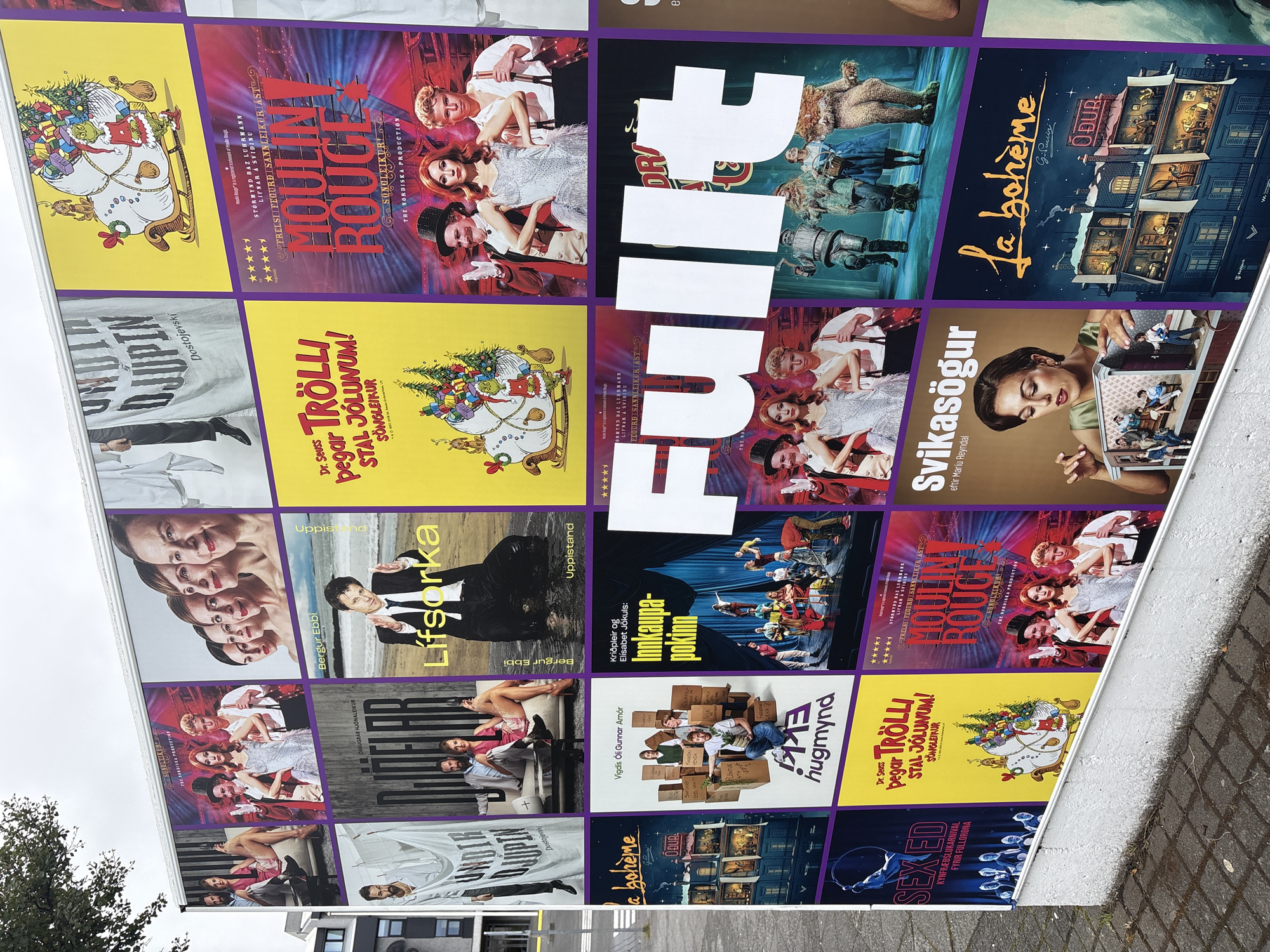 The image shows a large outdoor advertisement billboard in Reykjavik, Iceland, displaying a collage of theatrical posters. The billboard is positioned on a wall next to a street with a sidewalk and some vegetation. The posters promote various performances, including "La Bohème," "Moulin Rouge!", "Sex Ed," and what appears to be "Dr. Seuss' Grinch." Some posters feature actors in costume, while others use stylized illustrations or photographs of performers. The overall impression is a vibrant display of cultural events, likely at or near a theater in the city.