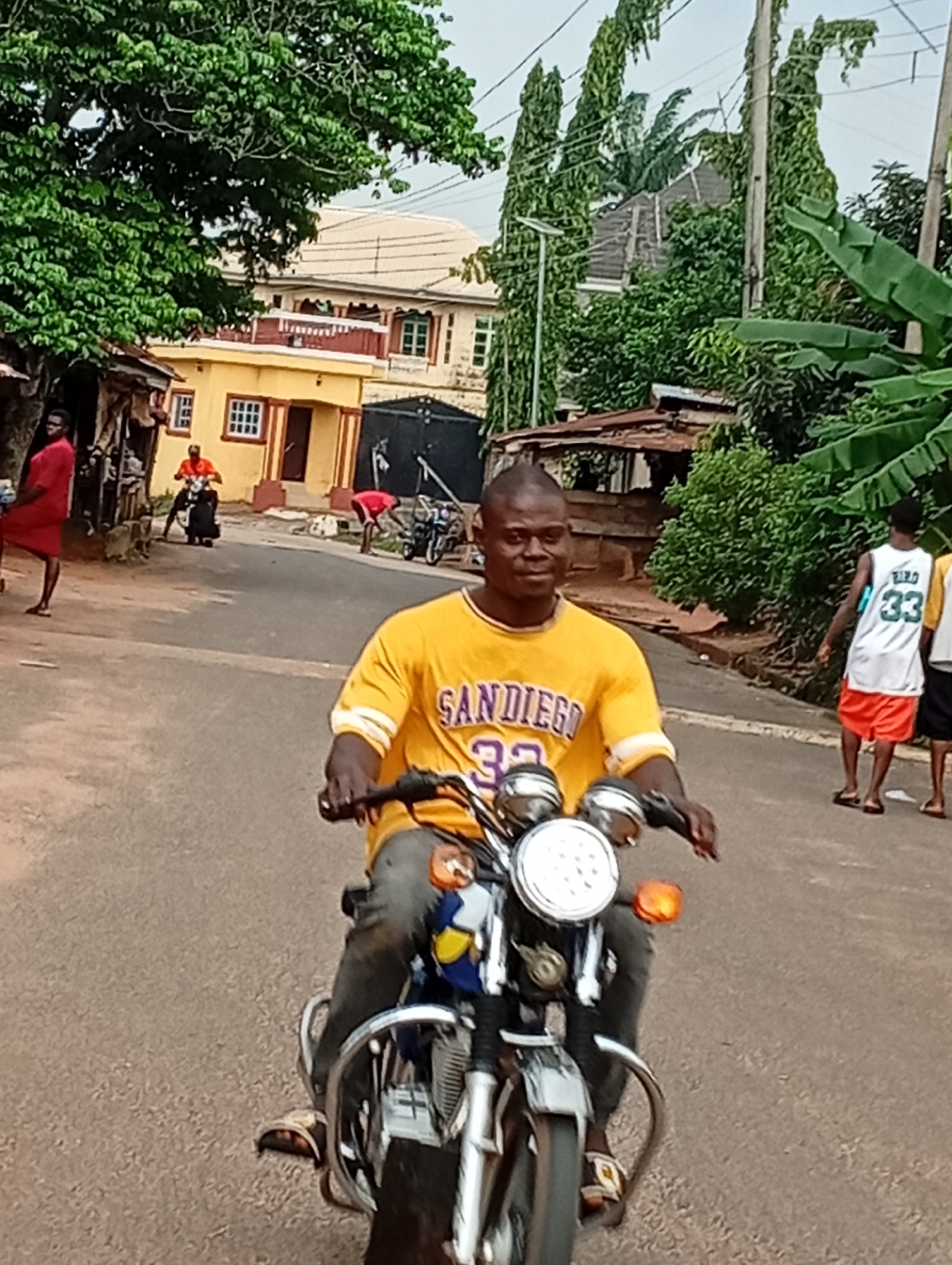 A man rides a motorcycle down a paved road in Nsugbe, Nigeria. He wears a yellow t-shirt with "San Diego" and the number "32" printed on it, along with grey pants. He is looking towards the camera and smiling. In the background, two other men stand on the side of the road. One wears a white tank top with "BIRD 33" printed on it and orange shorts, while the other wears a red shirt and black pants. Various buildings and lush green trees surround the road, indicating a rural or semi-urban setting. The sky is overcast.