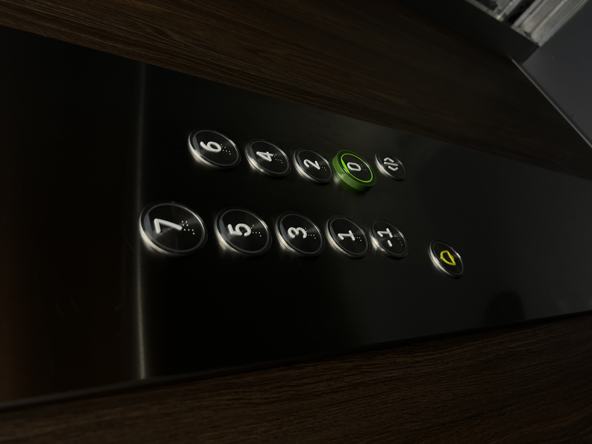 The image shows a close-up of an elevator panel. The panel is made of brushed metal and has a dark wood grain texture on the right side. There are several buttons on the panel, arranged in two columns. The left column has buttons labeled "7", "5", "3", "1", "-1", and a yellow alarm button. The right column has buttons labeled "6", "4", "2", "0", and a button with a "</>" symbol. The "0" button is illuminated green, indicating the elevator is currently on the ground floor. The lighting is dim, creating reflections on the metal surface. There are no people visible in the image. The context of Florești, Romania, is not visually represented in this image.